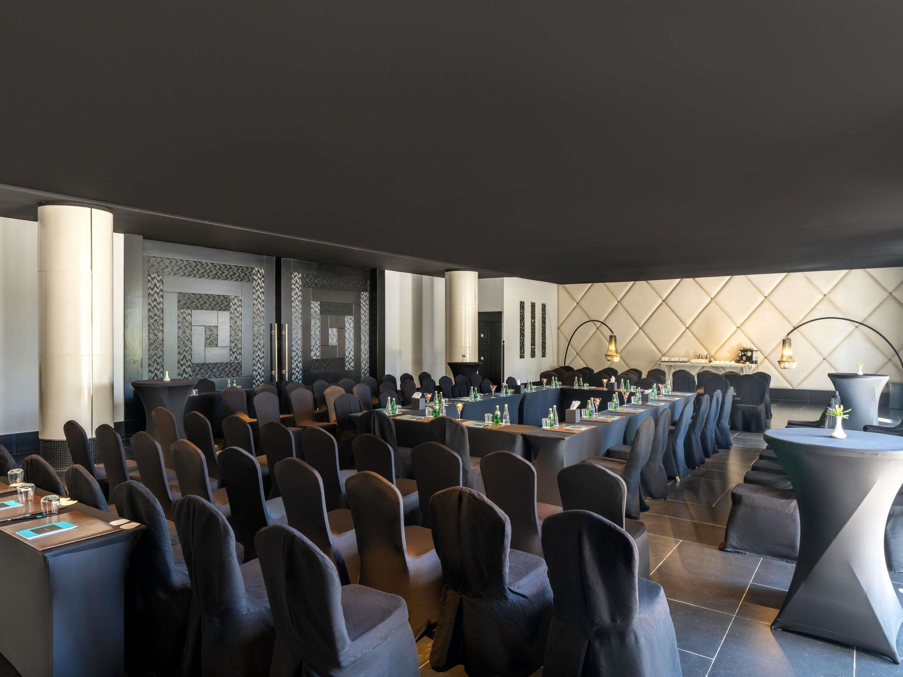 Un salón de conferencias con varias mesas y sillas, un diseño de pared moderno y una planta grande en maceta.