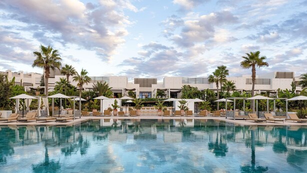 Piscina con sillas de descanso y sombrillas, rodeada de palmeras y edificios modernos.