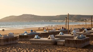Un área de descanso frente a la playa con mobiliario y cojines de madera, con vistas al mar y una colina distante con escritura árabe.