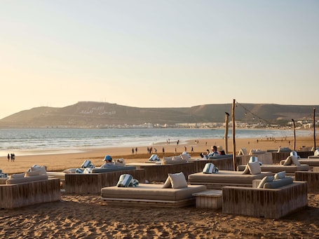 A beachside lounge area with wooden furniture and cushions, overlooking the sea and a distant hill with Arabic script.