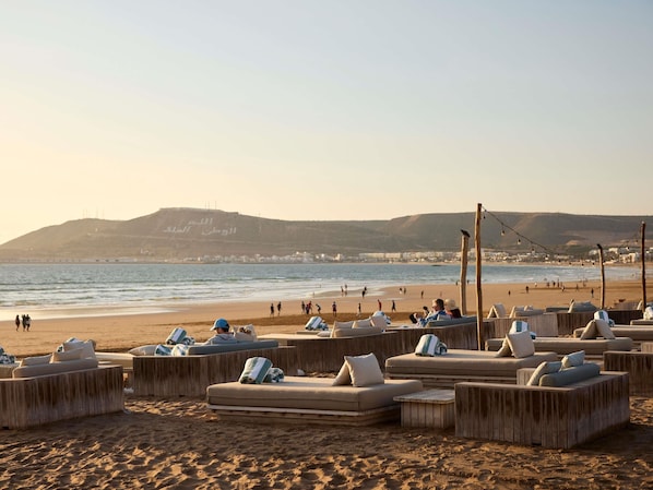A beachside lounge area with wooden furniture and cushions, overlooking the sea and a distant hill with Arabic script. - Sofitel Agadir Thalassa Sea & Spa (Agadir)