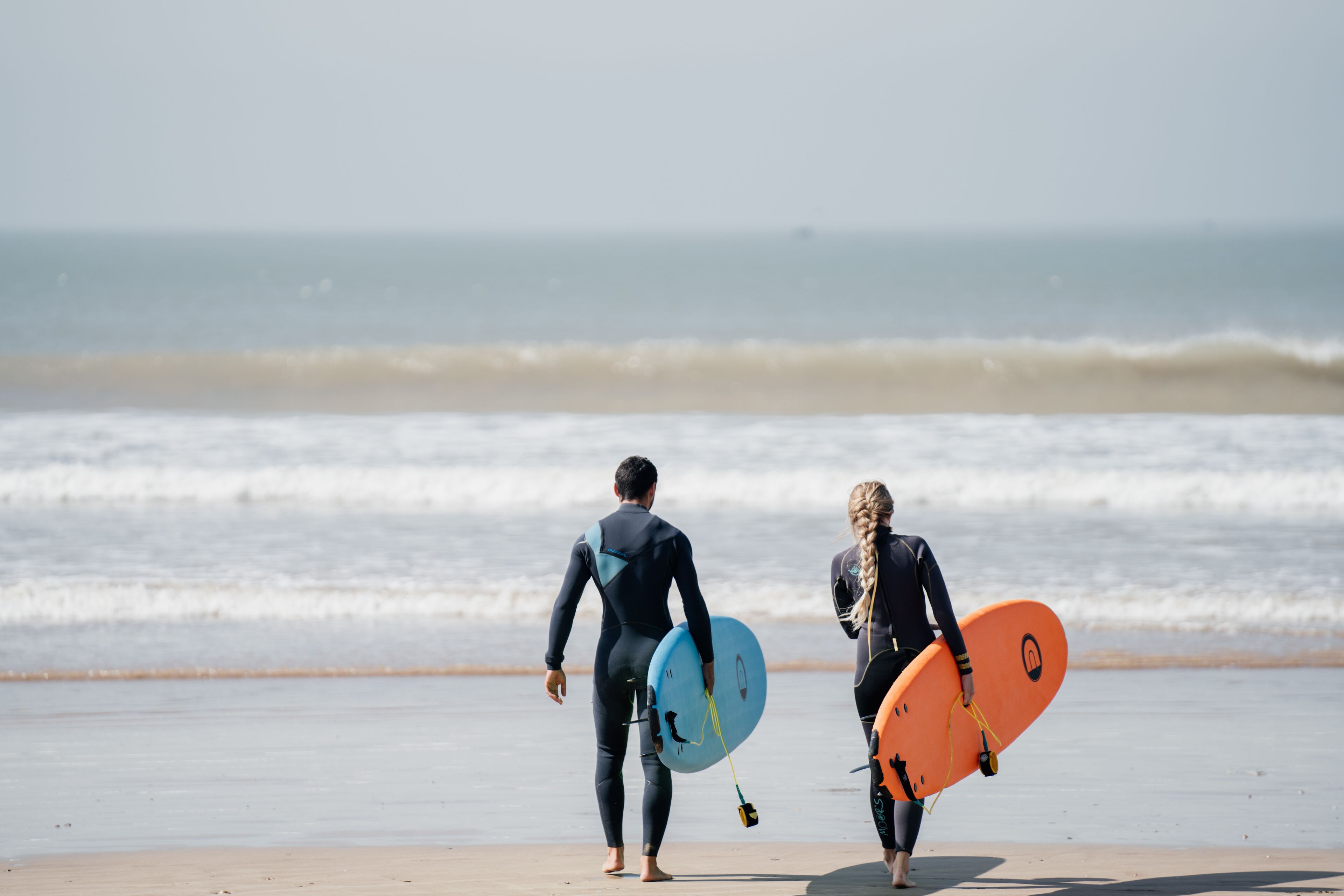 Two individuals walking on a beach with surfboards.