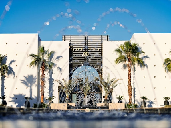 A modern building with a large fountain, palm trees, and a clear sky.