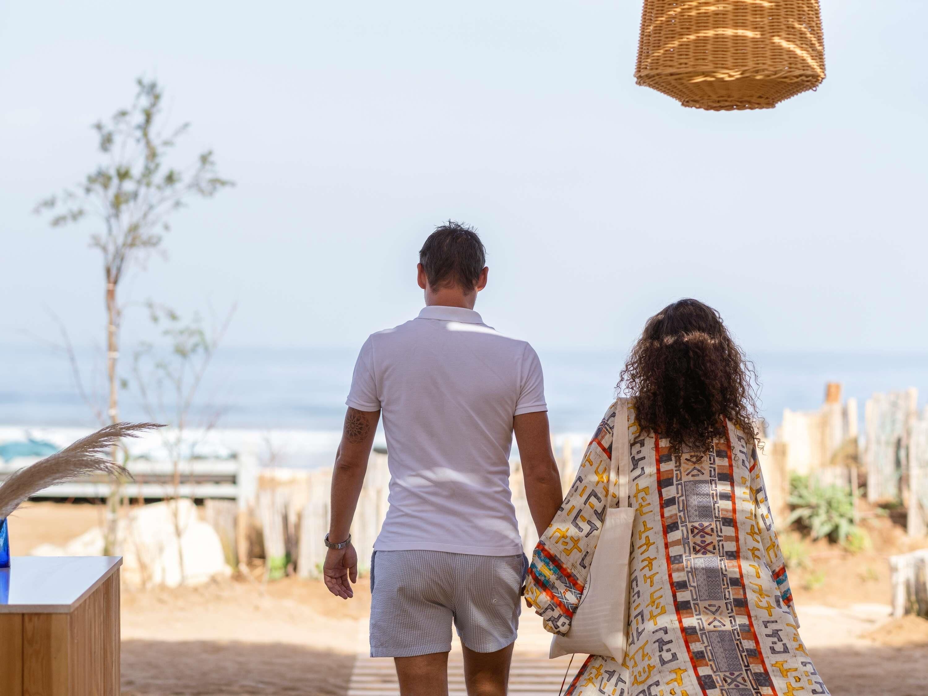 A couple walking on a wooden deck overlooking a beach with a clear view of the ocean and a city skyline in the distance.