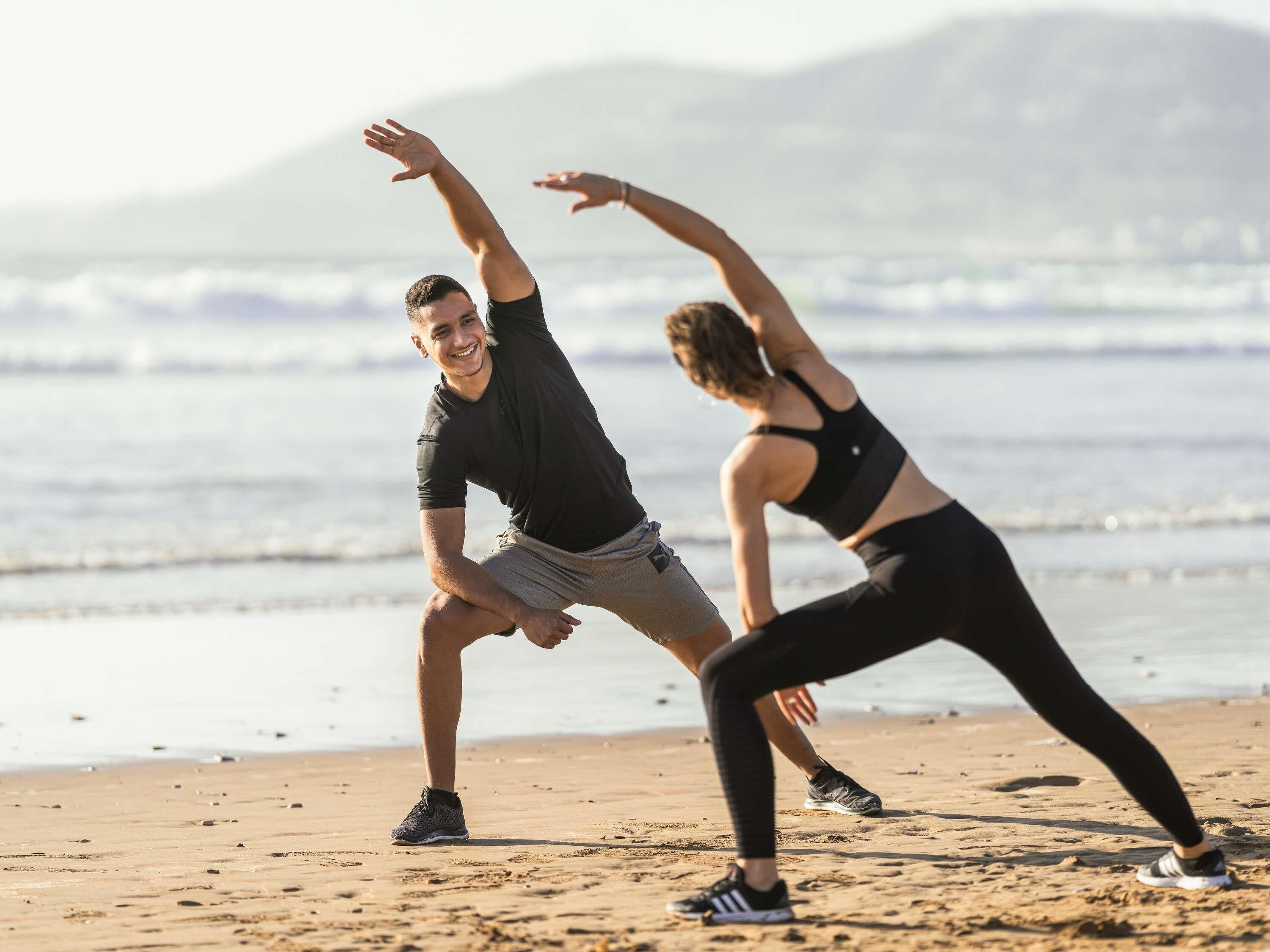 Two people stretching on a beach with a mountain in the background.