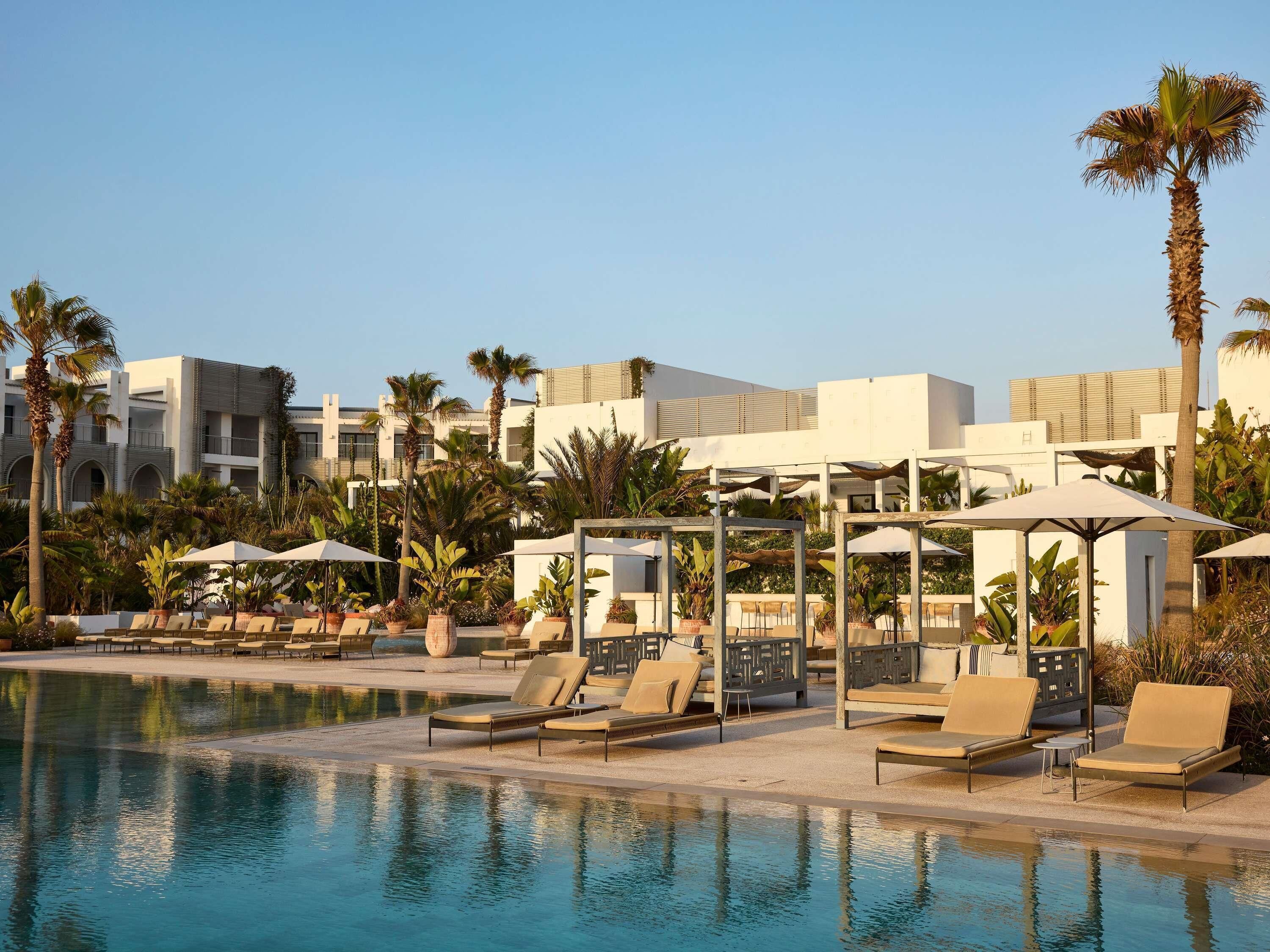 A poolside area with lounge chairs and umbrellas, surrounded by palm trees and modern buildings.