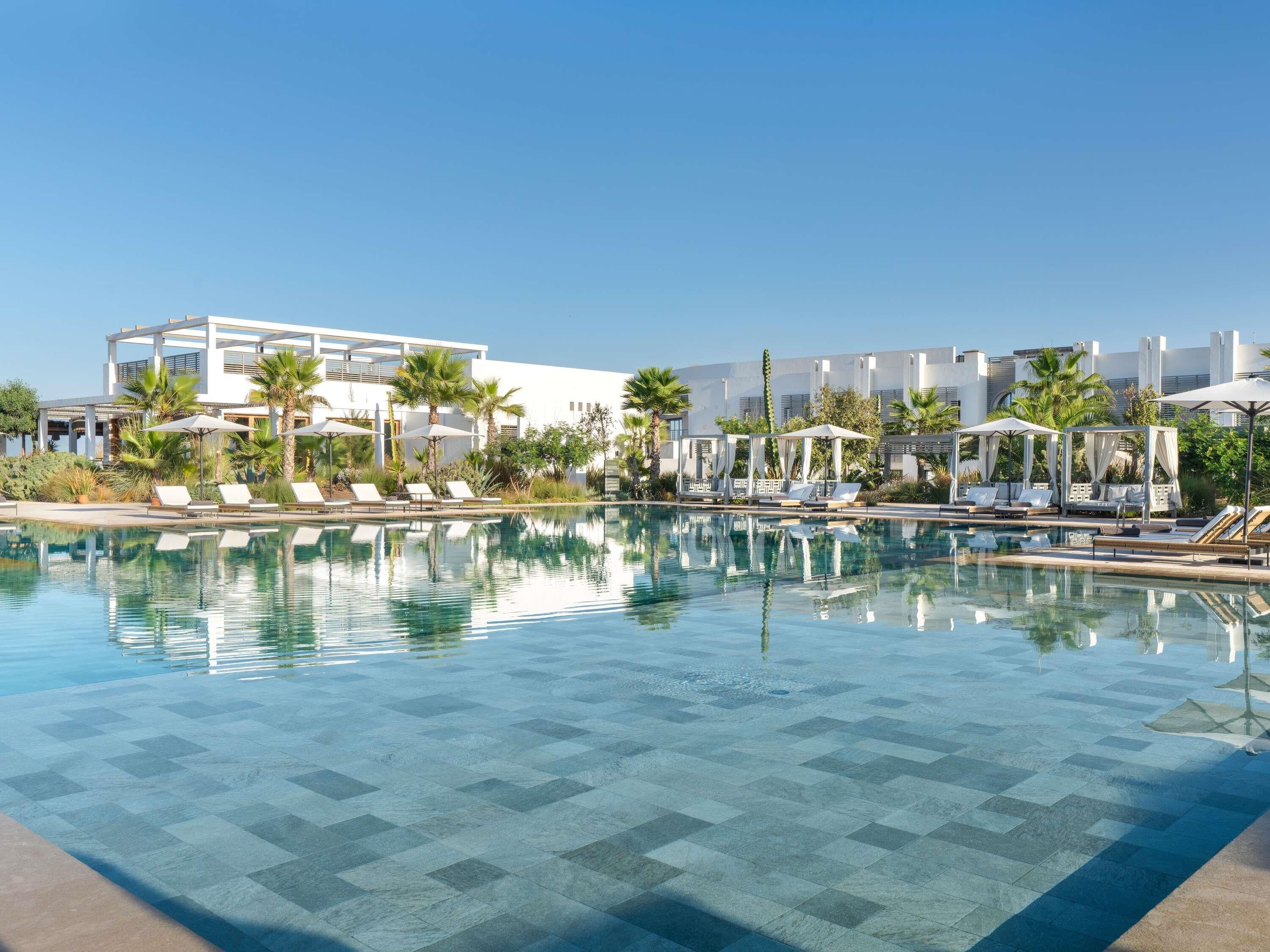 Un área de piscina con sillas de descanso y sombrillas, rodeada de palmeras y edificios modernos.