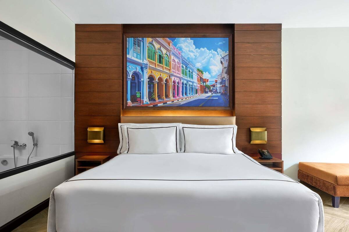 A hotel room with a large bed, a painting of a colorful street scene, a bathtub, and two bedside lamps.