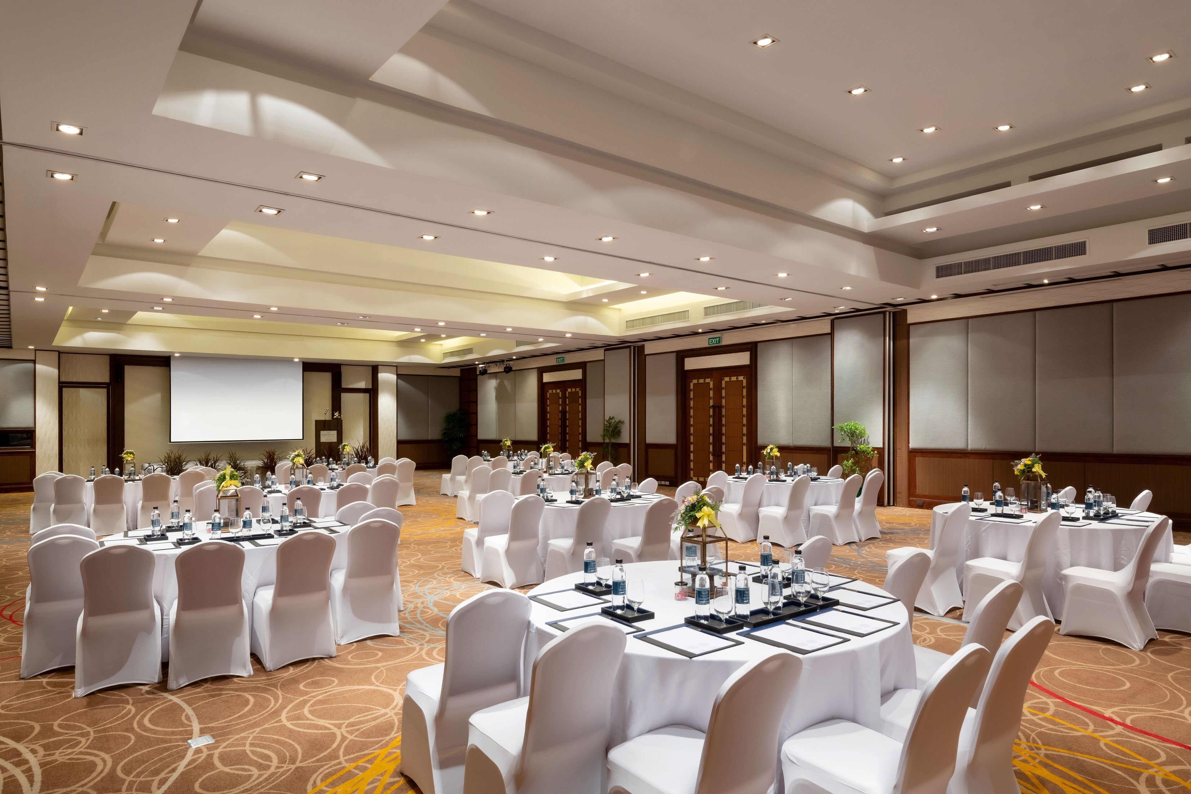 Een ruime congreszaal met meerdere ronde tafels, witte stoelen en bloemstukken als decoratie.