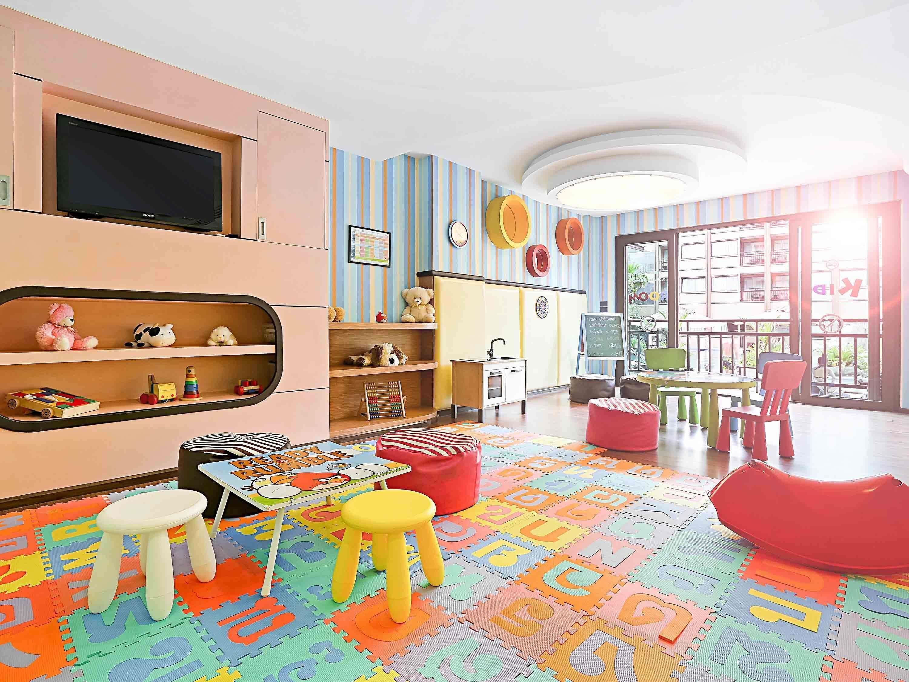 A children's playroom with a television, shelves with toys, a colorful play mat, and a view of the outside.