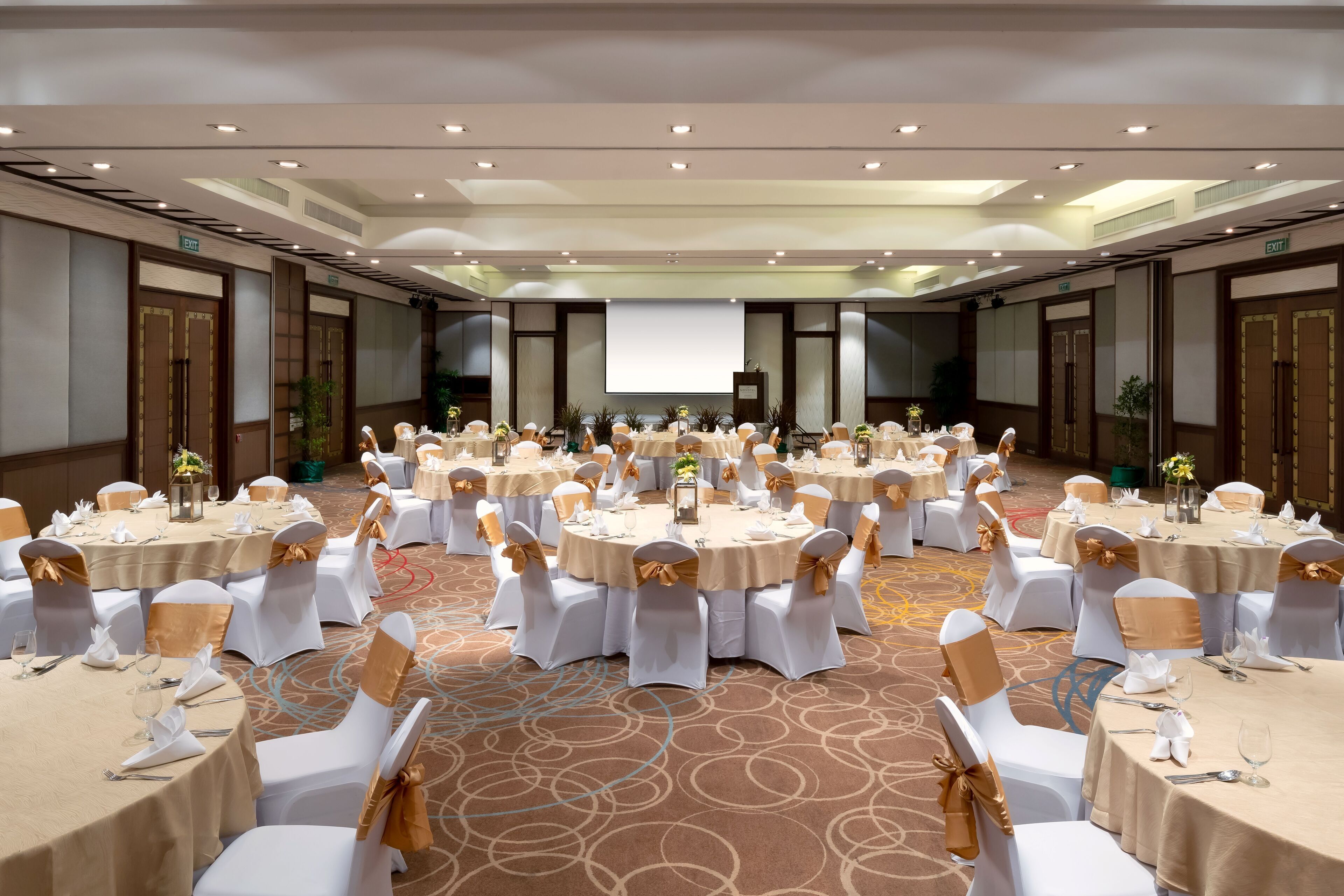 A large conference room with multiple round tables, each set with white chairs and gold sashes, and a carpeted floor.