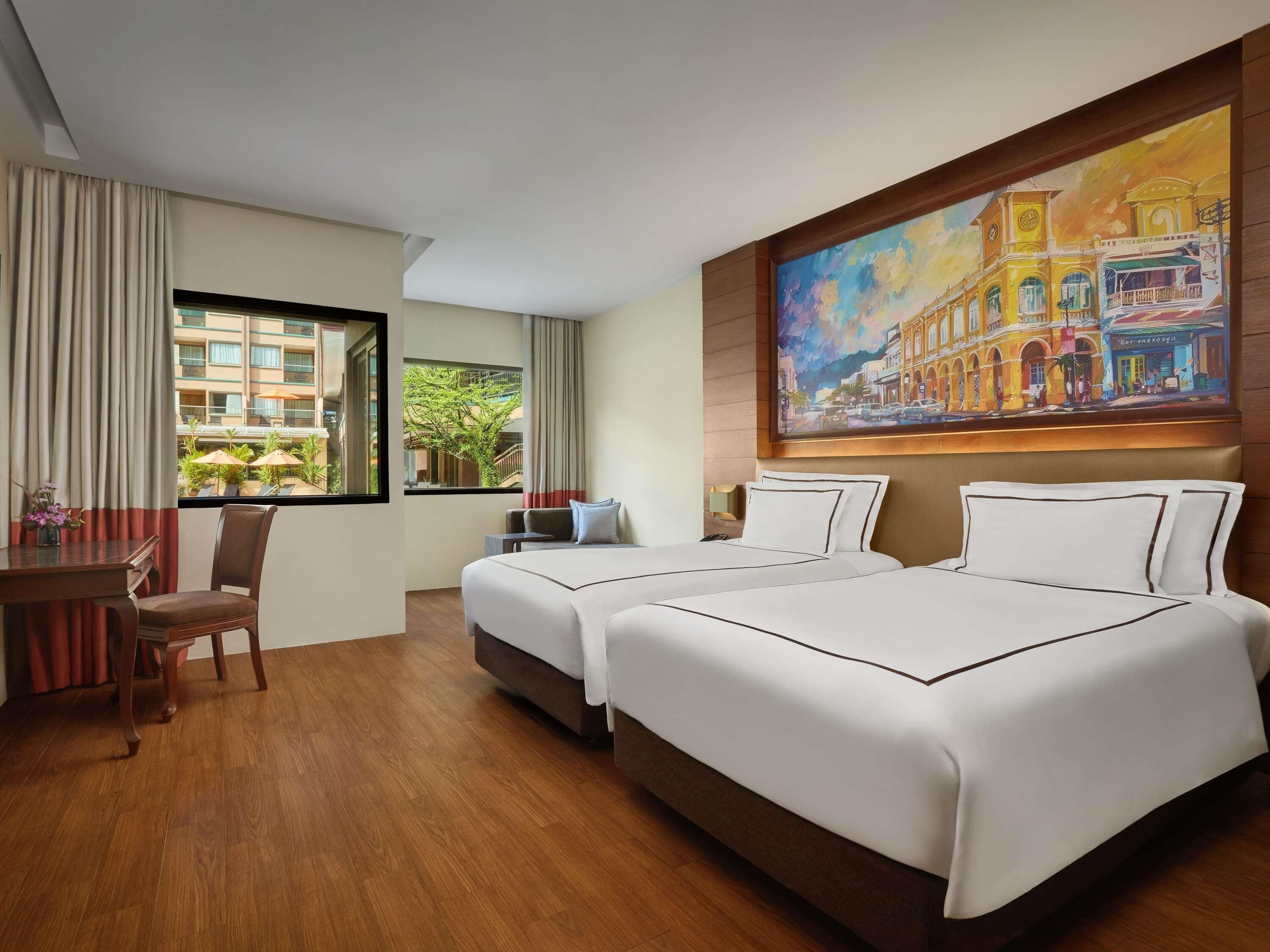 A hotel room with two beds, a desk, and a chair. There is a large window with a view of buildings and greenery outside.