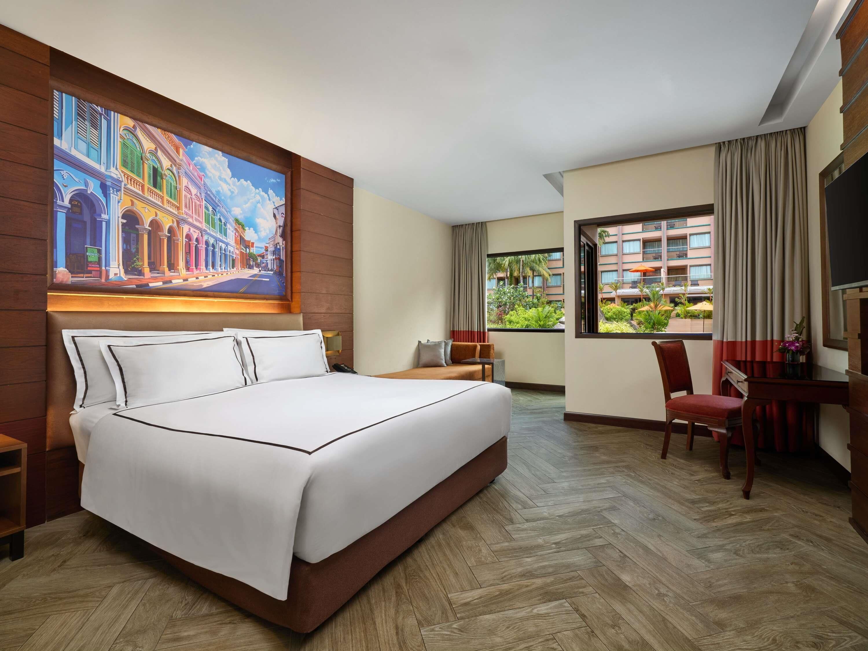 Une chambre d’hôtel avec un grand lit, un bureau, une chaise, un tableau au mur et une vue sur un immeuble et la verdure environnante.