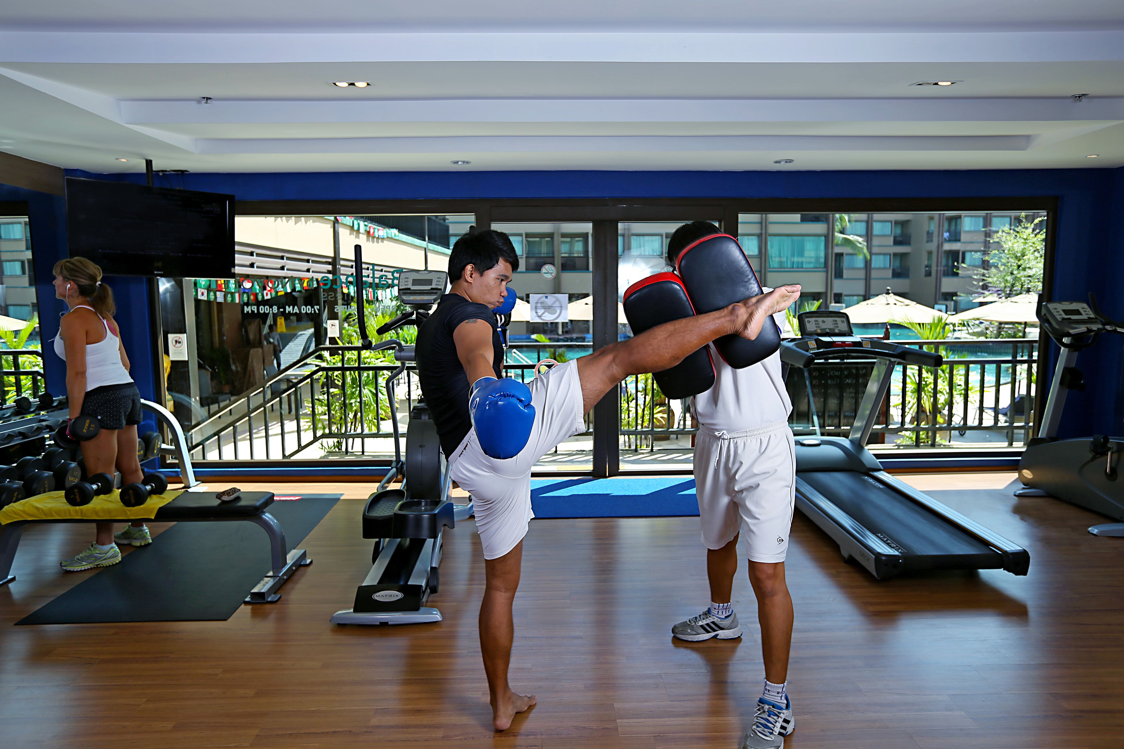 Een sportschool met trainingsapparatuur, een loopband en uitzicht op een zwembad en gebouwen.