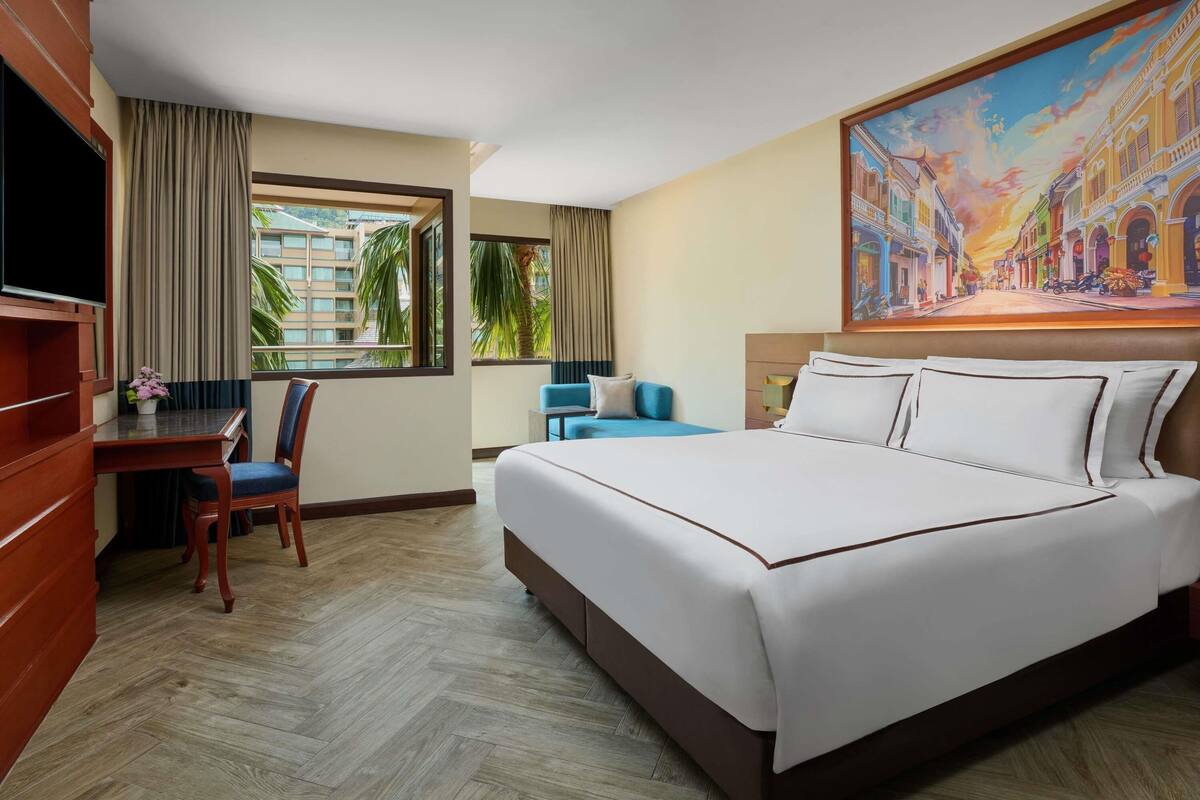 A hotel room with a bed, a desk, a chair, a TV, and a painting on the wall.