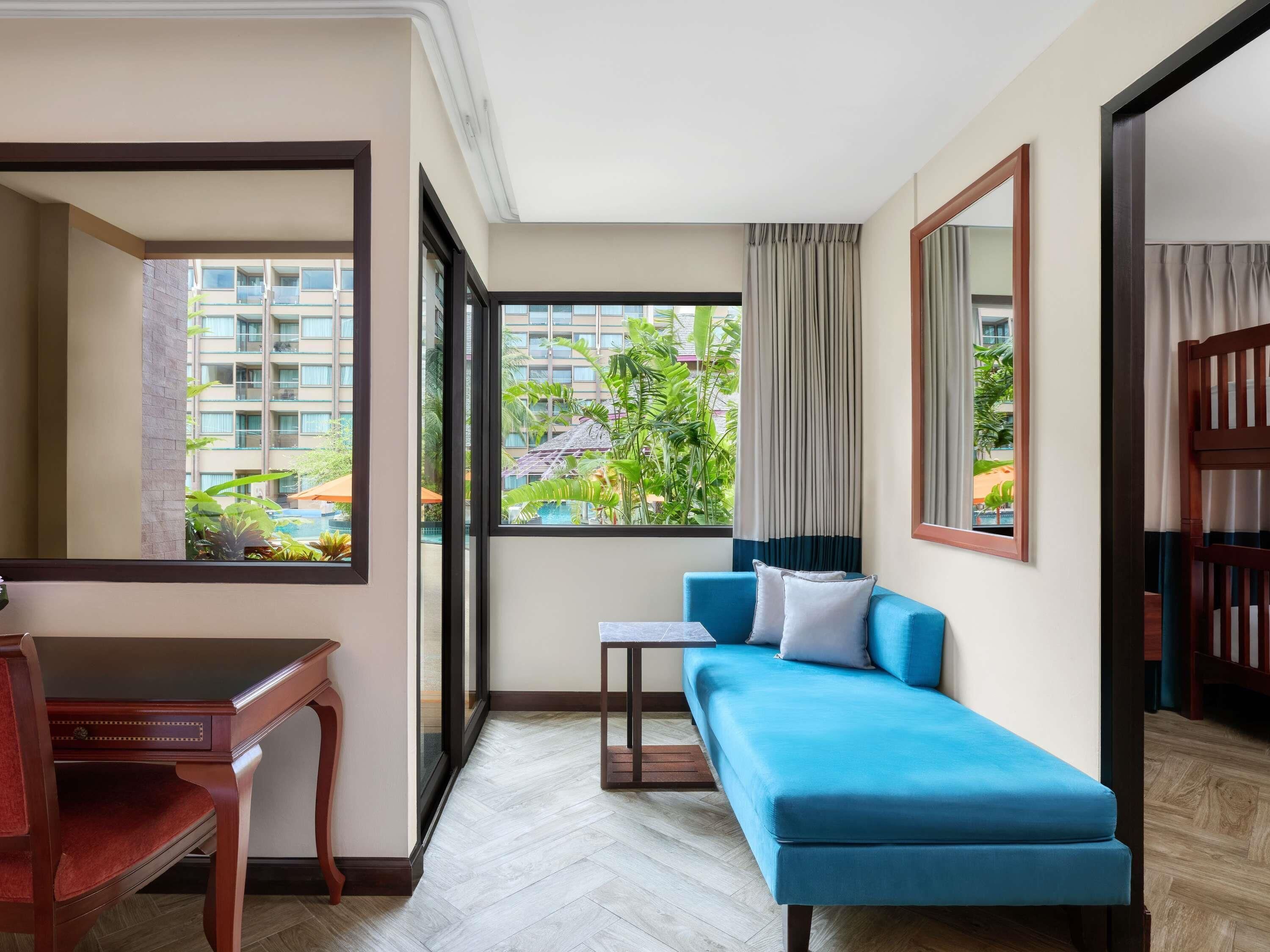 A hotel room with a sofa, a desk, and a large window with a view of a building and greenery.