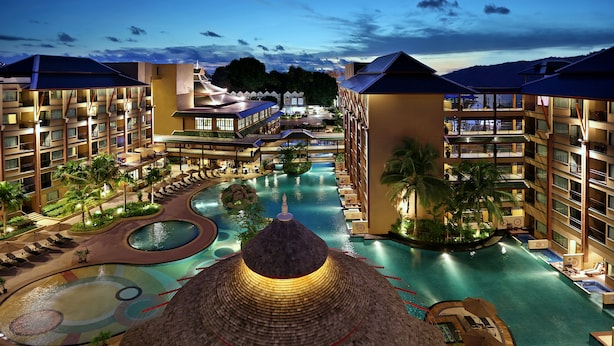 Un resort con varias piscinas, un elemento central de agua y un amplio pabellón con techo de paja.