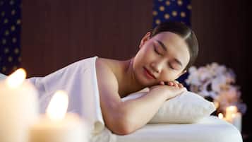A person lying on a bed with a white pillow, surrounded by candles and a floral arrangement.