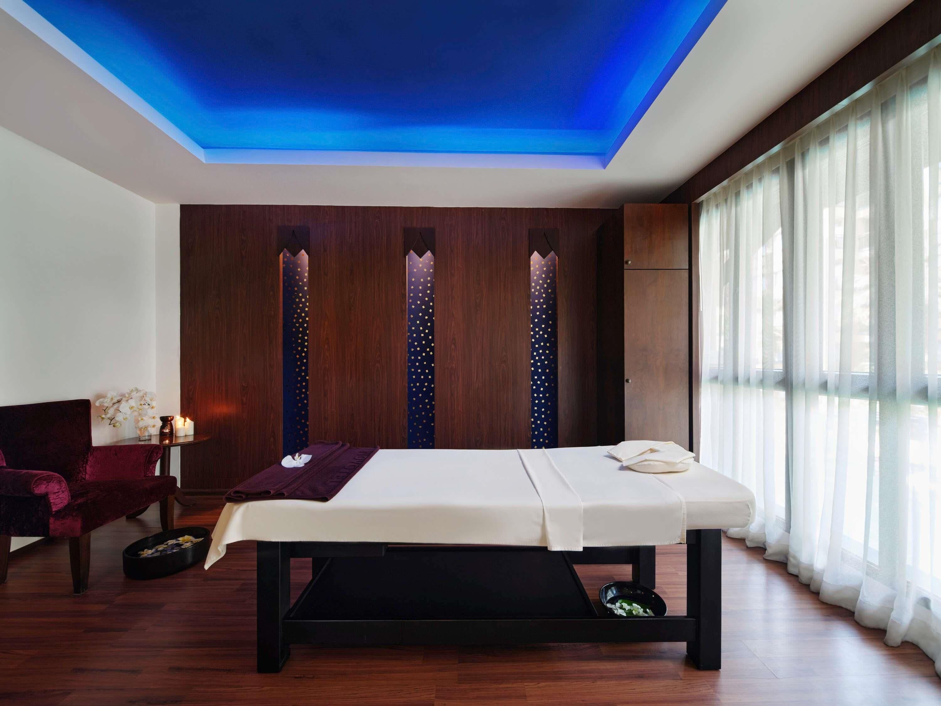 Quarto com mesa de massagem, paredes de painel de madeira, uma cadeira vermelha e janela com cortinas leves.