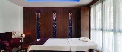 Quarto com mesa de massagem, paredes de painel de madeira, uma cadeira vermelha e janela com cortinas leves.