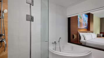 Un baño moderno con ducha envuelta en vidrio, una bañera independiente y un espejo grande que refleja un dormitorio con cama y un cuadro.