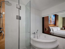 A modern bathroom with a glass-enclosed shower, a freestanding bathtub, and a large mirror reflecting a bedroom with a bed and a painting.