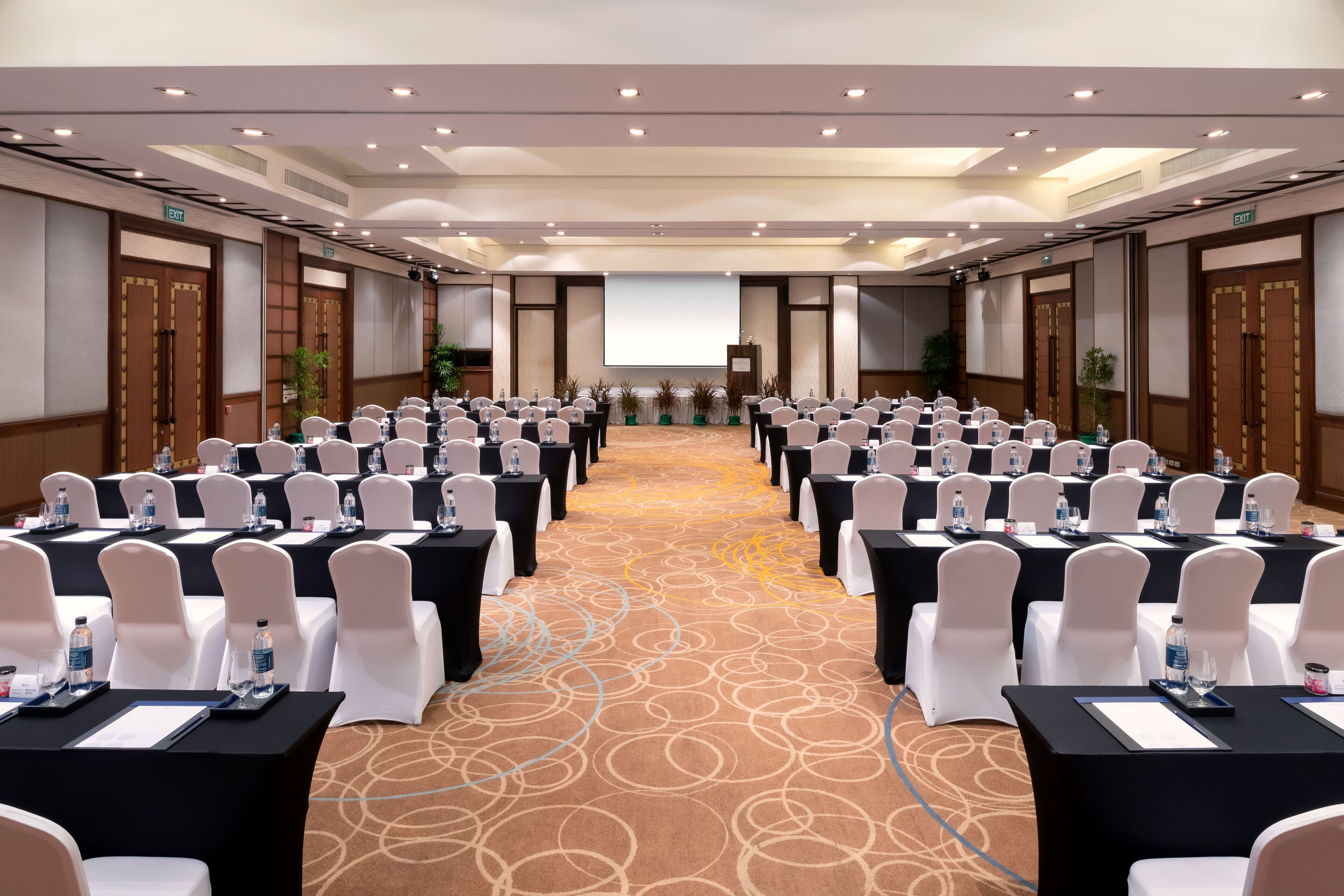 A conference room with rows of tables and chairs, a projector screen at the front, and an exit sign on the wall.