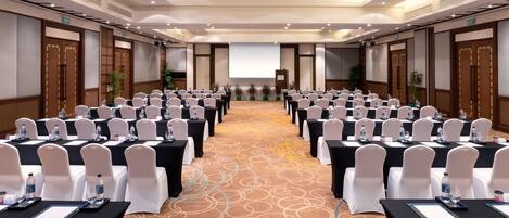 A conference room with rows of tables and chairs, a projector screen at the front, and an exit sign on the wall.