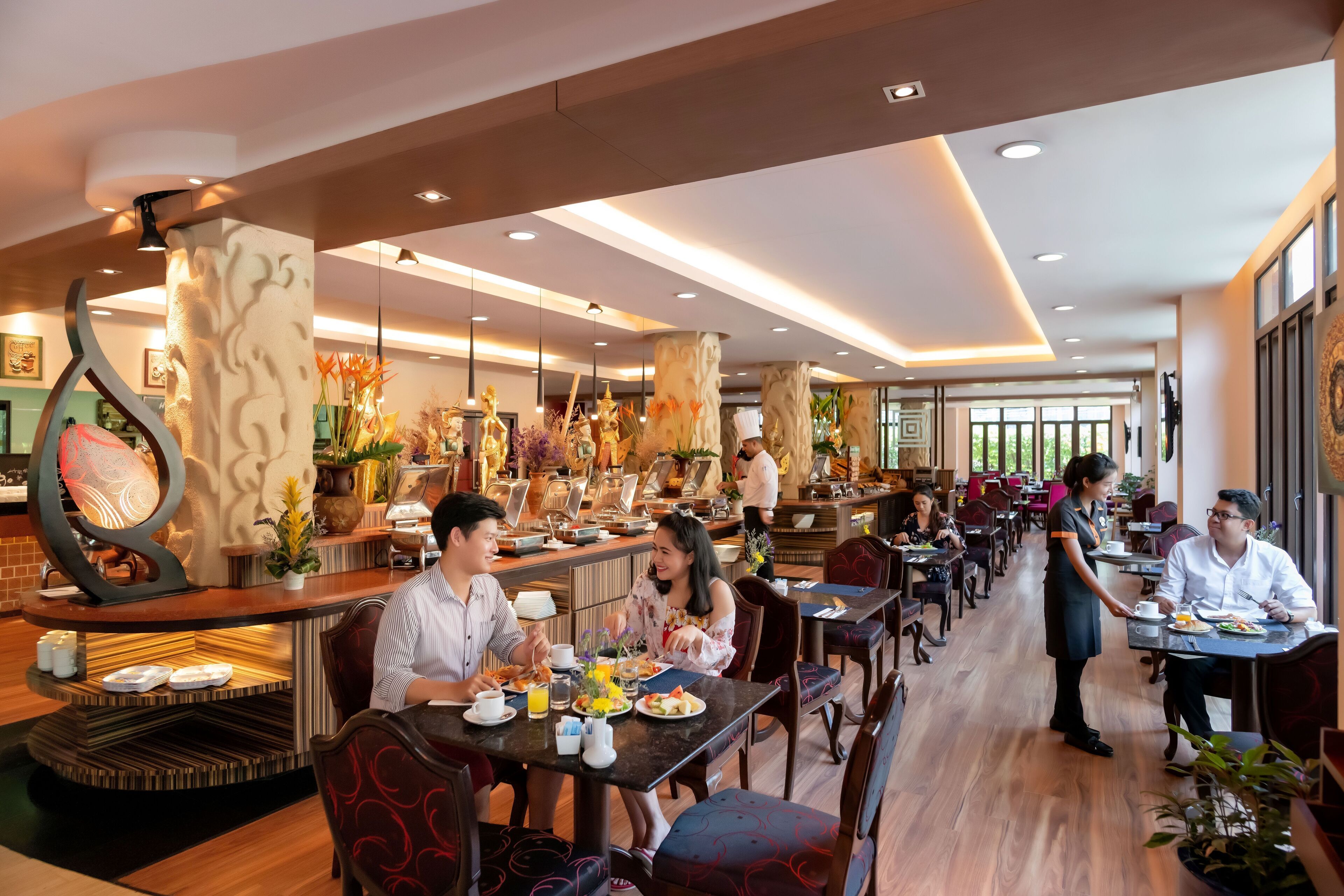 A modern dining area with wooden flooring, tables, and chairs. There are decorative elements like plants and a unique wall sculpture.