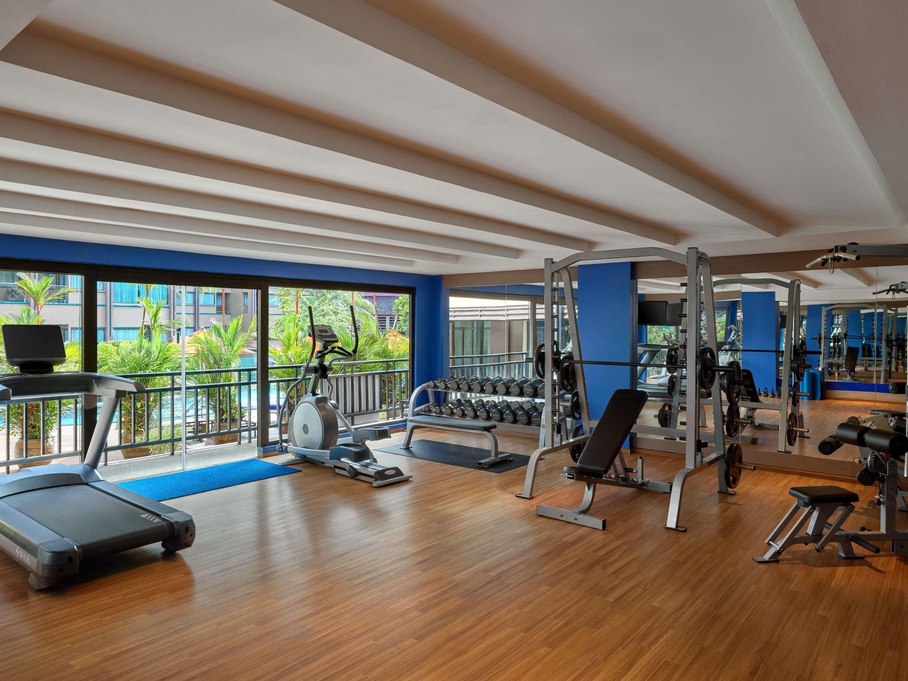 Une salle de sport équipée de matériel d’exercice, d’un tapis de course, et offrant une vue sur une piscine et un espace verdoyant à l’extérieur.