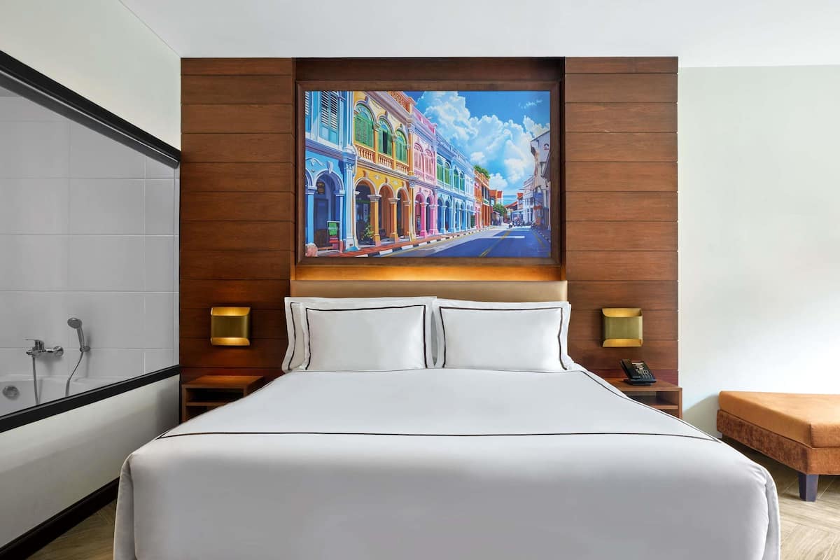 A hotel room with a large bed, a painting of a colorful street scene, a bathtub, and two bedside lamps.