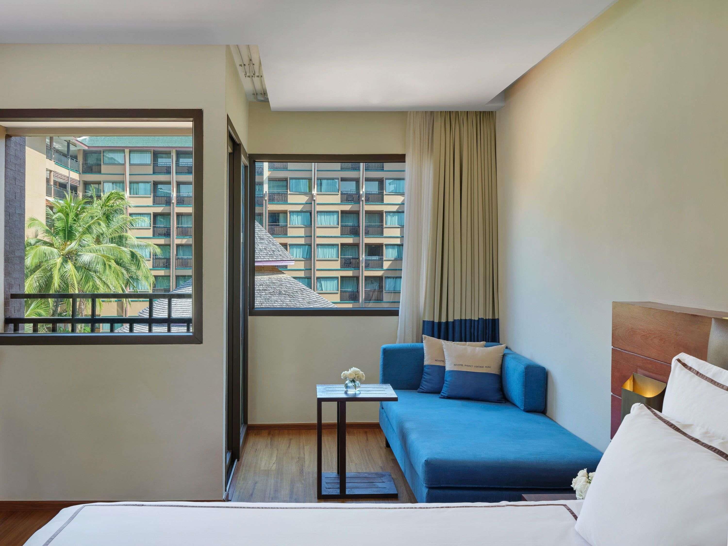 A hotel room with a bed, a blue sofa, a small table with a vase, a window with a view of a building and palm trees, and a curtain.