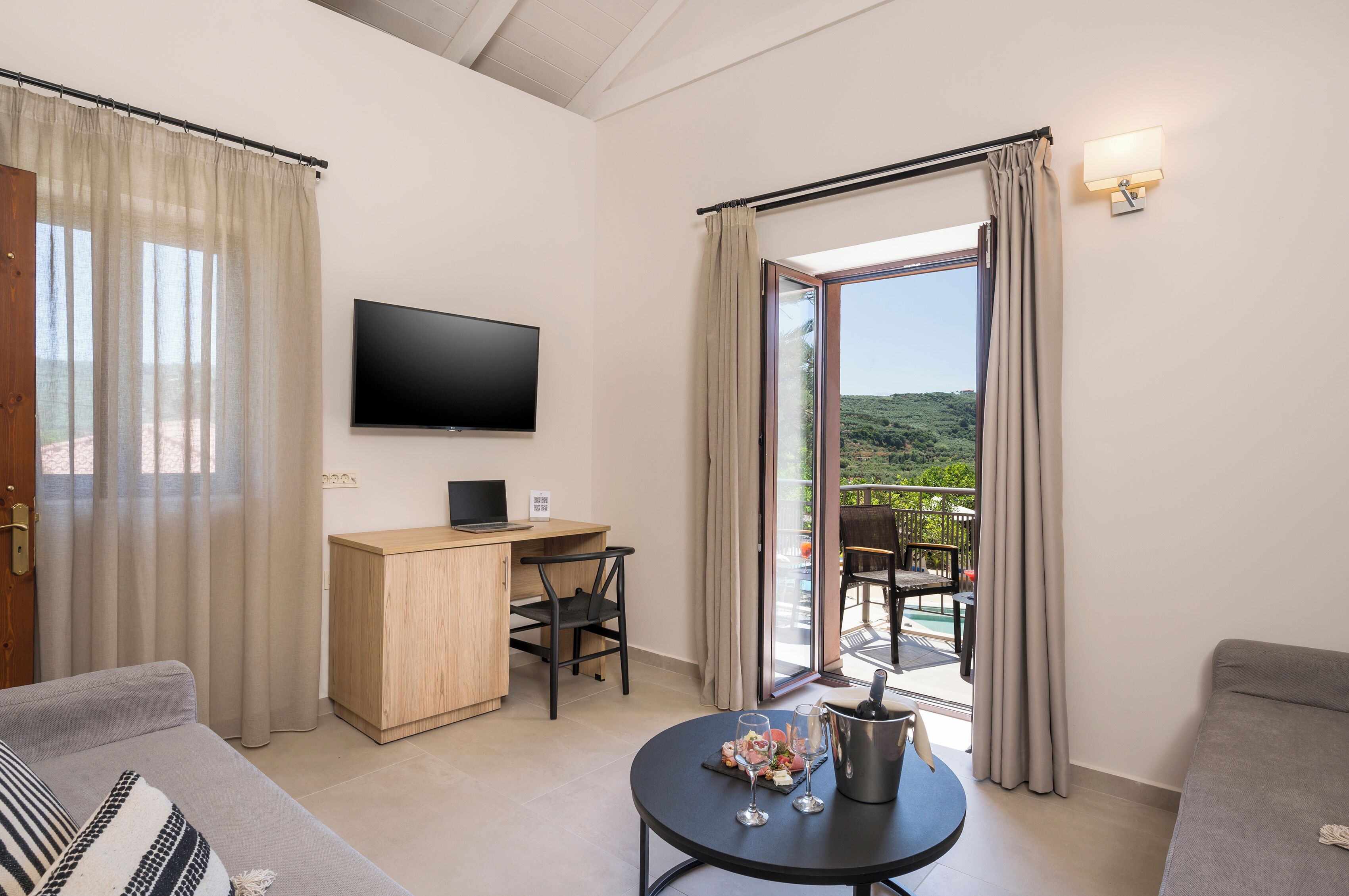Quarto moderno com TV de tela plana, escrivaninha de madeira, mesa de jantar com garrafa e copos, sofá e varanda com vista para o verde.