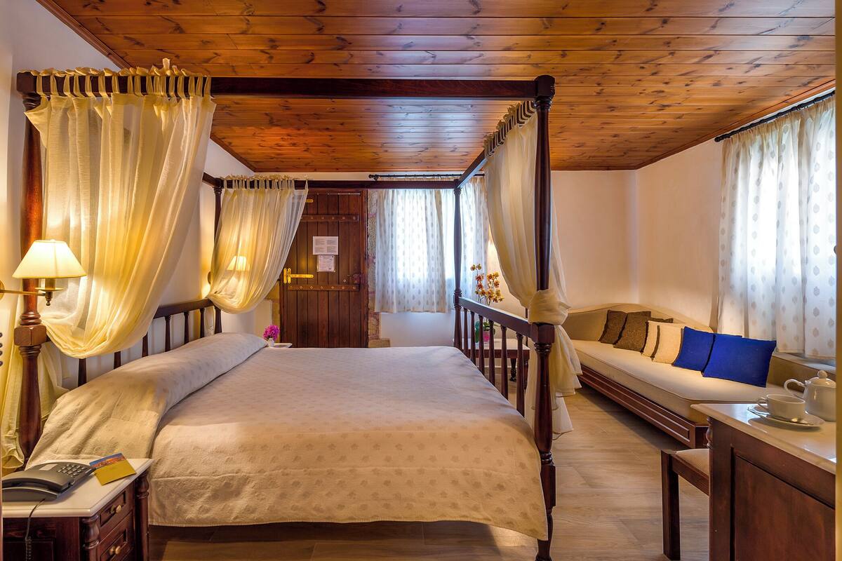 A four-poster bed with a canopy, a sofa, a bedside table with a lamp, and a wooden desk with a teapot and cup.