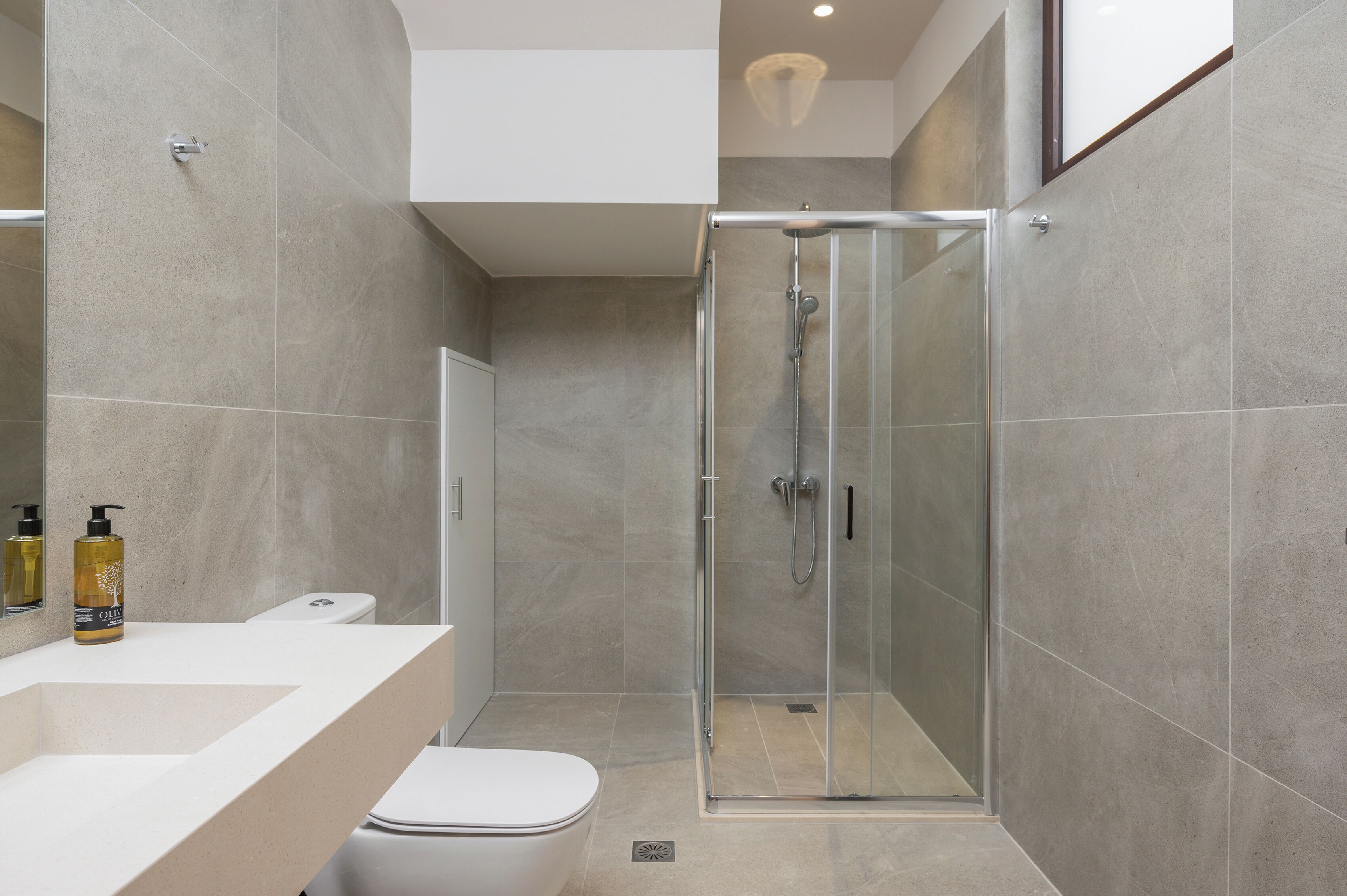 Un baño moderno con ducha de vidrio, inodoro blanco y encimera color crema con artículos de aseo.