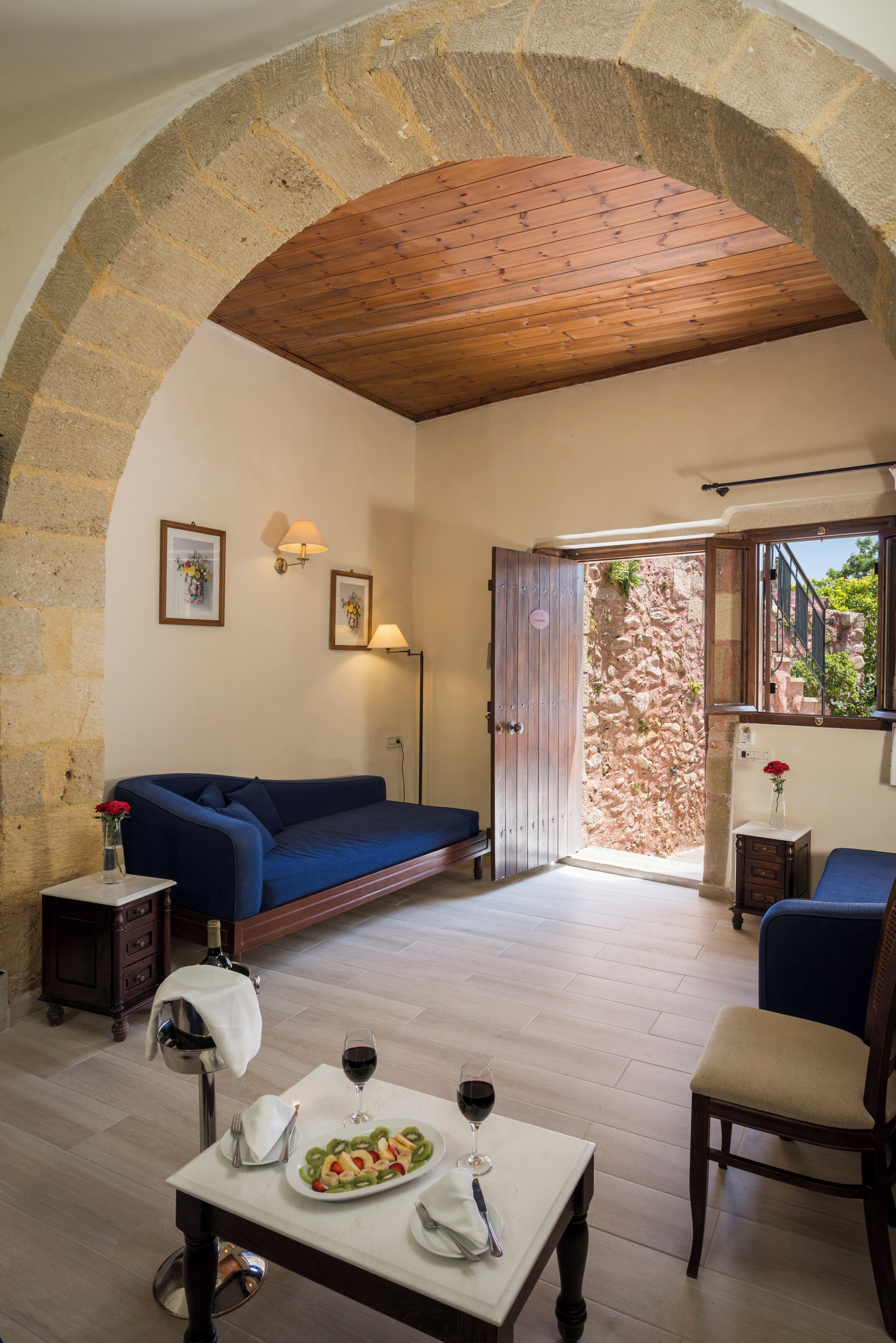 Una habitación acogedora con techo de madera, paredes de piedra, un sofá azul, una mesa de comedor con comida y vino, y vistas a una pared de piedra y follaje exterior.