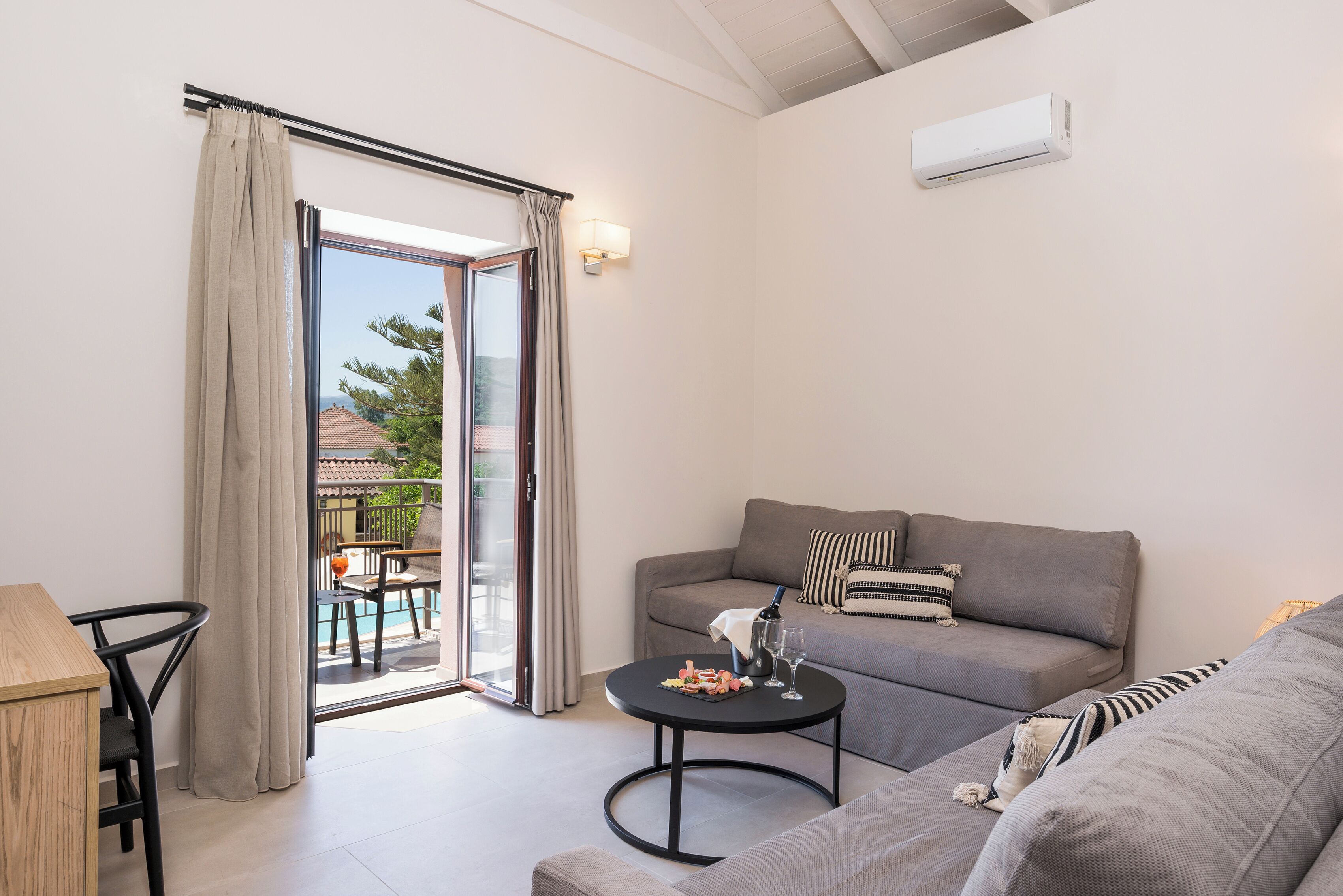 A modern living room with a grey sofa, a round coffee table, and a dining area with a wooden table and chairs. A sliding glass door leads to an outdoor area with a pool and a view of a building with a tiled roof.