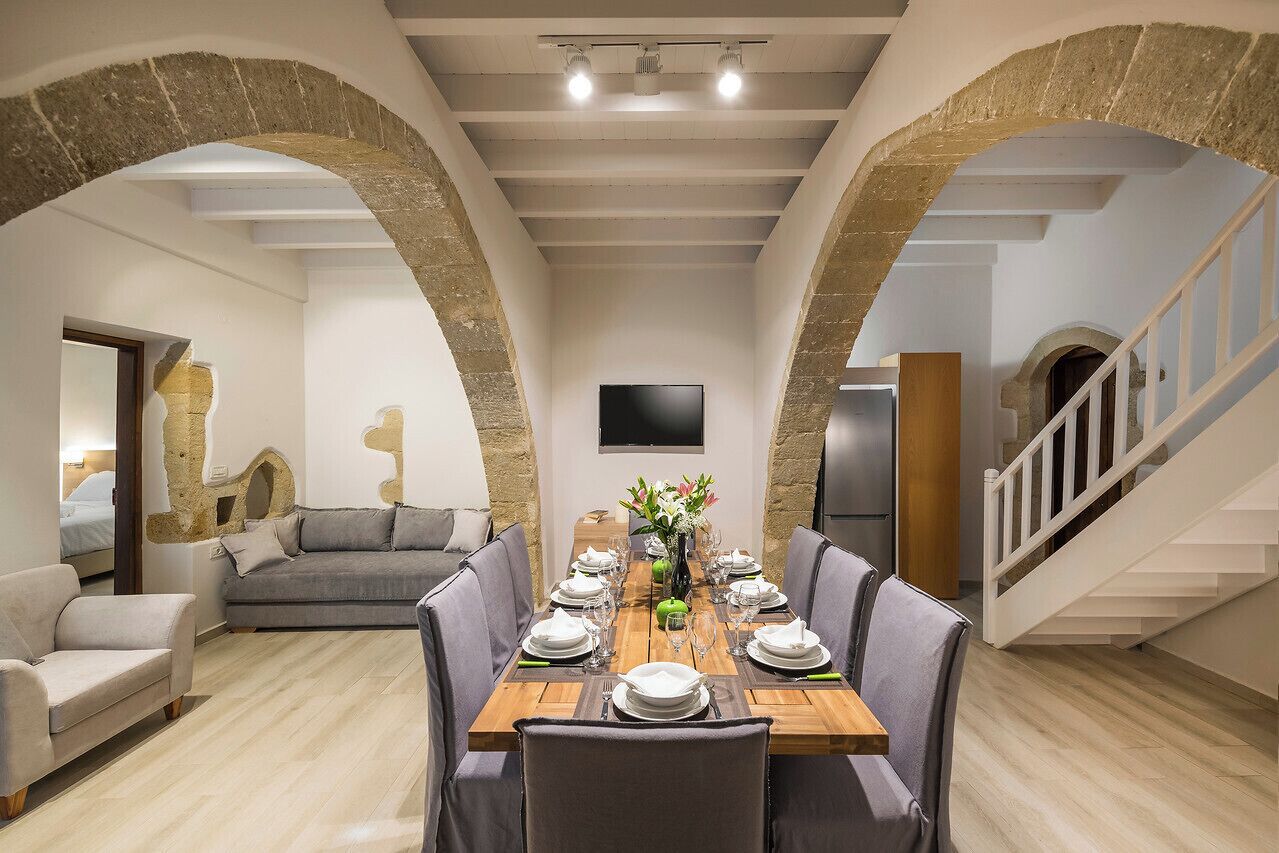 Un coin repas avec une table en bois dressée pour six personnes, bordé d’arches en pierre, d’un canapé gris et d’un escalier.