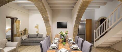 A dining area with a wooden table set for six, flanked by stone arches, a grey sofa, and a staircase.