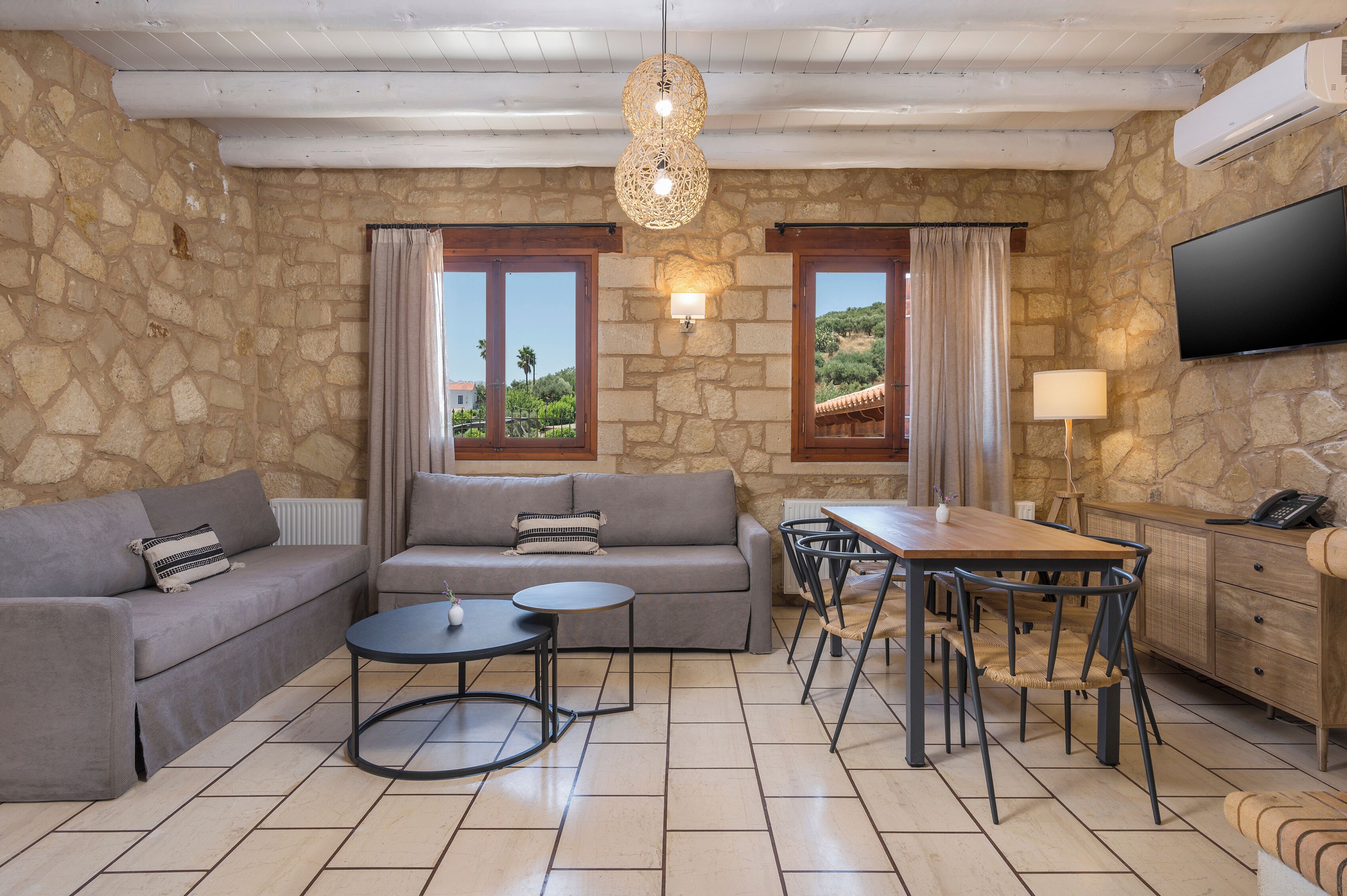 A living room with stone walls, a grey sofa, a dining table with chairs, a TV, and a lamp.