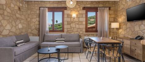 A living room with stone walls, a grey sofa, a dining table with chairs, a TV, and a lamp.