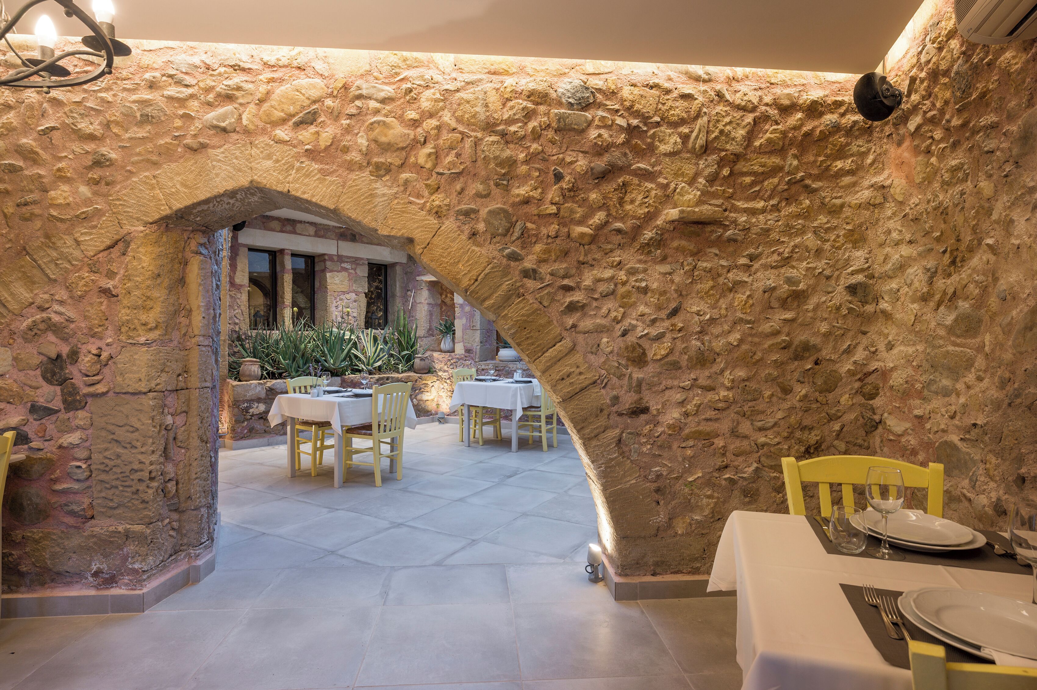 An interior dining area with stone walls, tables set for two, and a view into a garden.