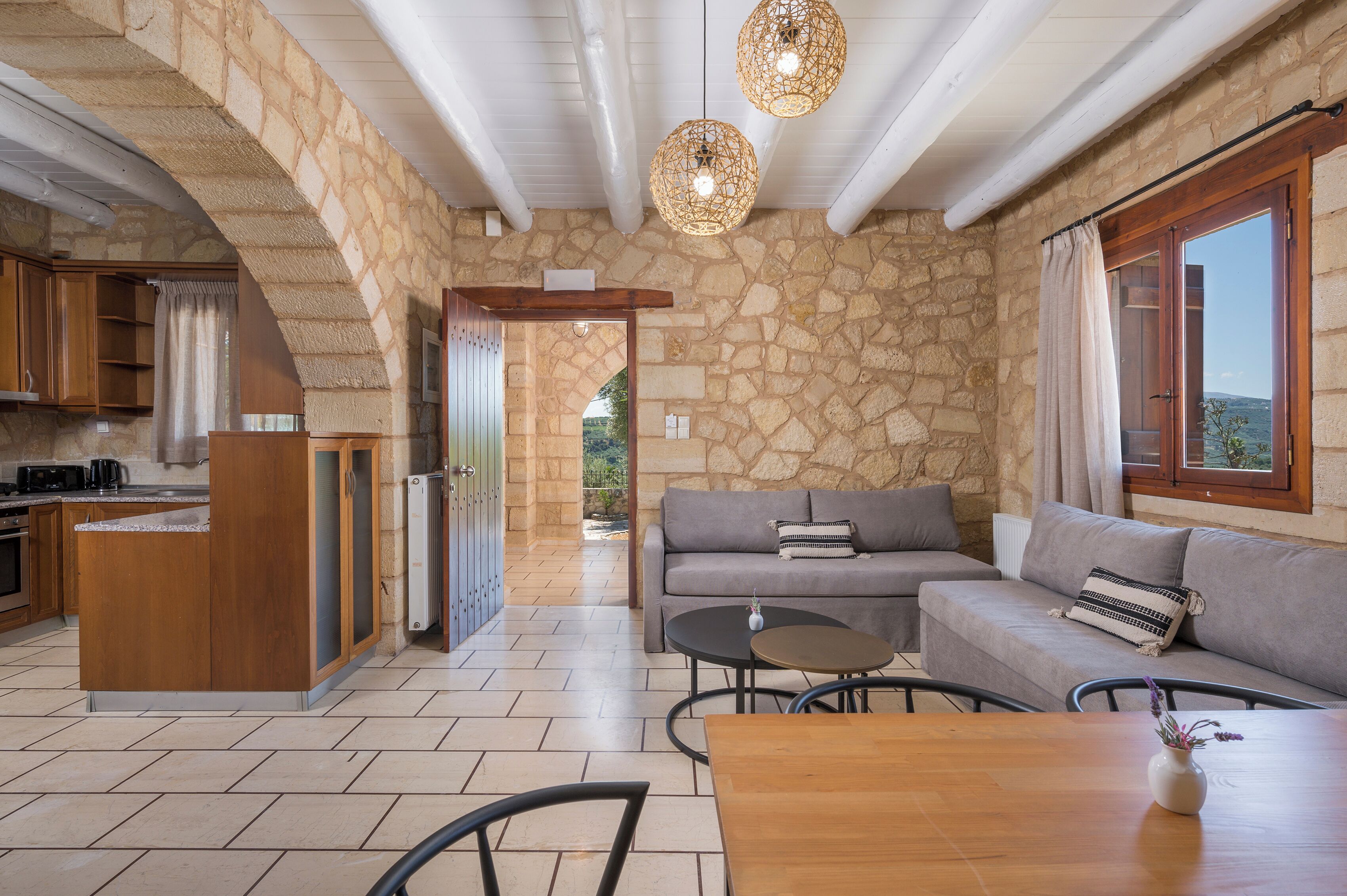 A spacious living area with a stone wall, a grey sofa, a wooden dining table, and a kitchen area with wooden cabinets and a built-in island.