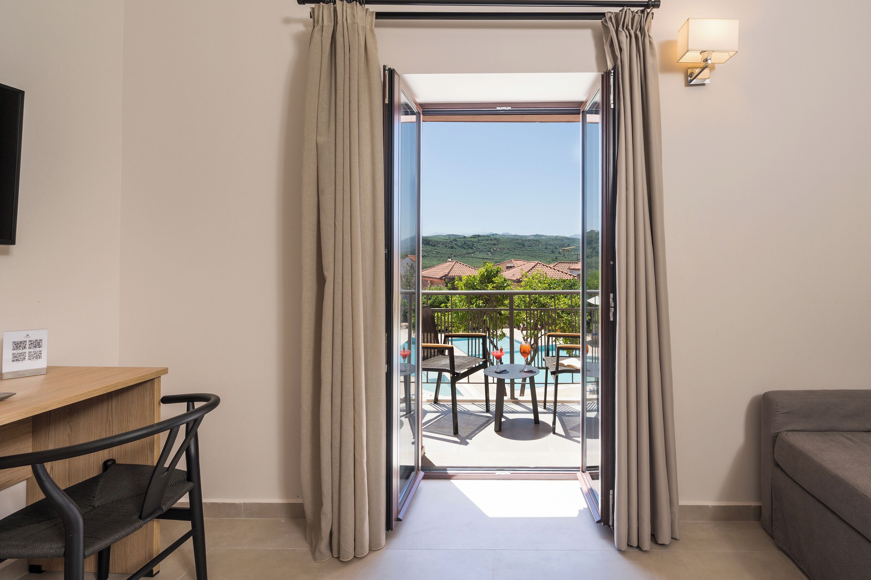 A hotel room with a desk, a chair, a sofa, and a balcony with a pool and outdoor furniture.