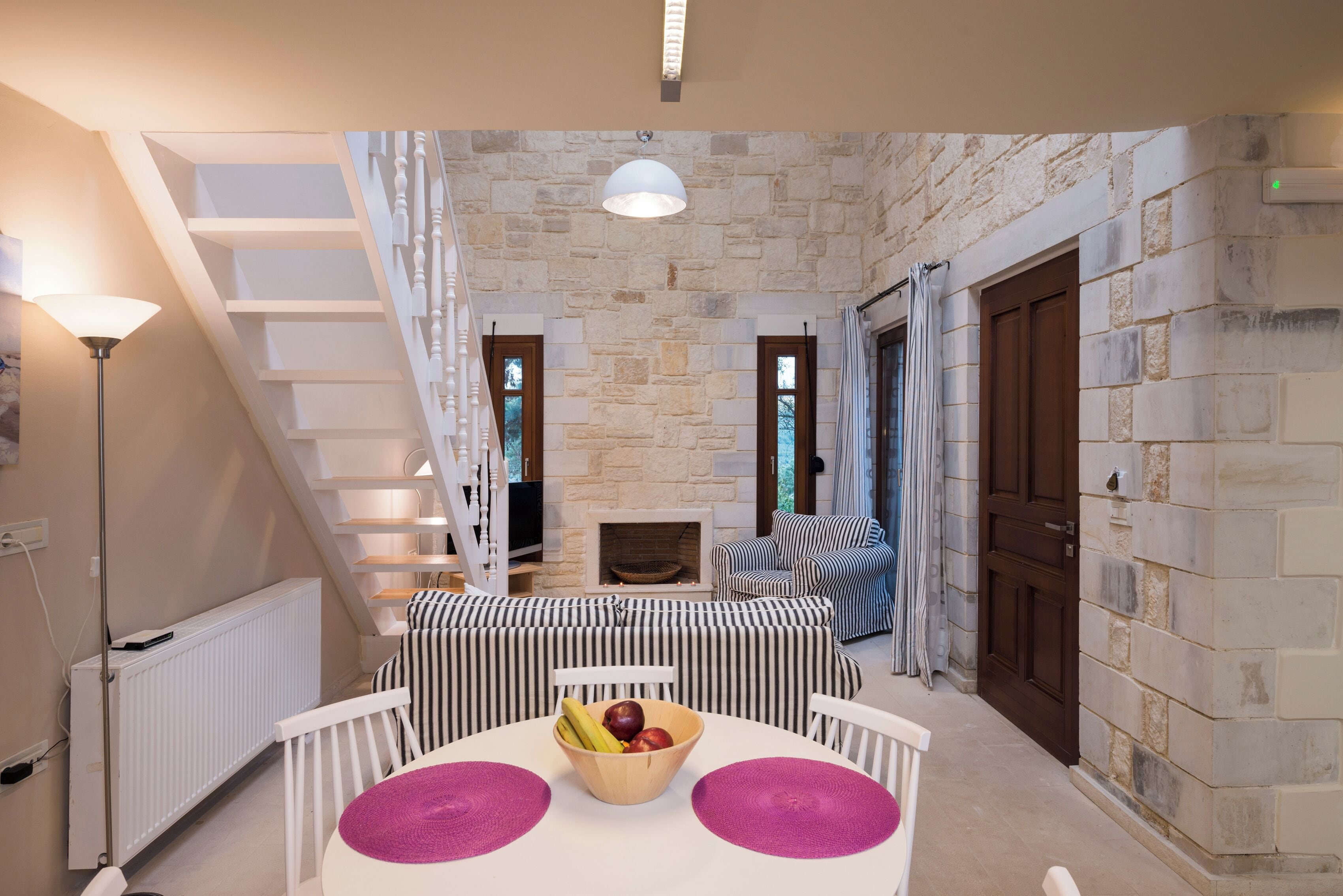 Una habitación acogedora con chimenea de piedra, sofá a rayas y una mesa de comedor con fruteros.