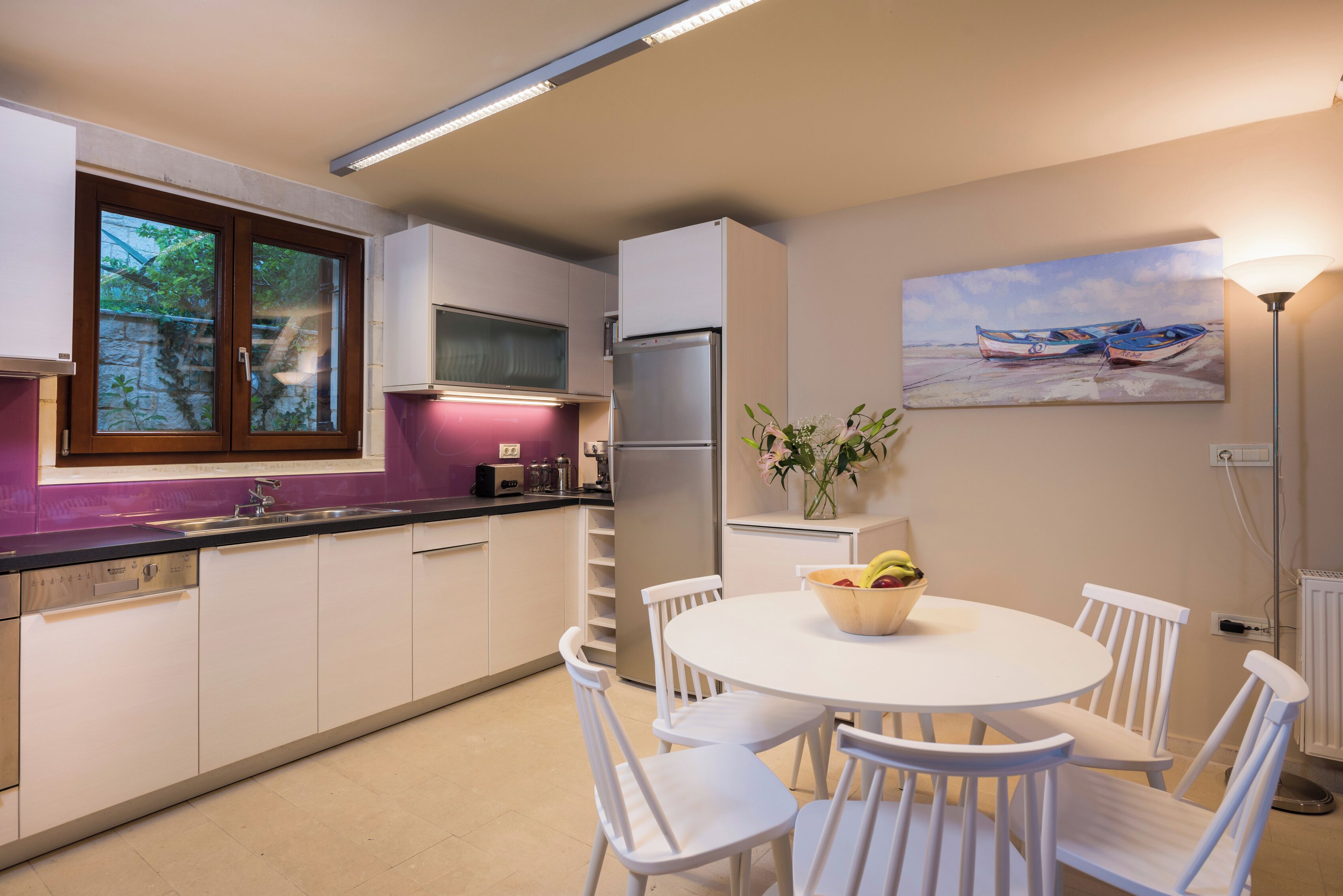 Een moderne keuken met witte kastjes, een roestvrijstalen koelkast, een eettafel met witte stoelen en een schilderij van boten aan de muur.