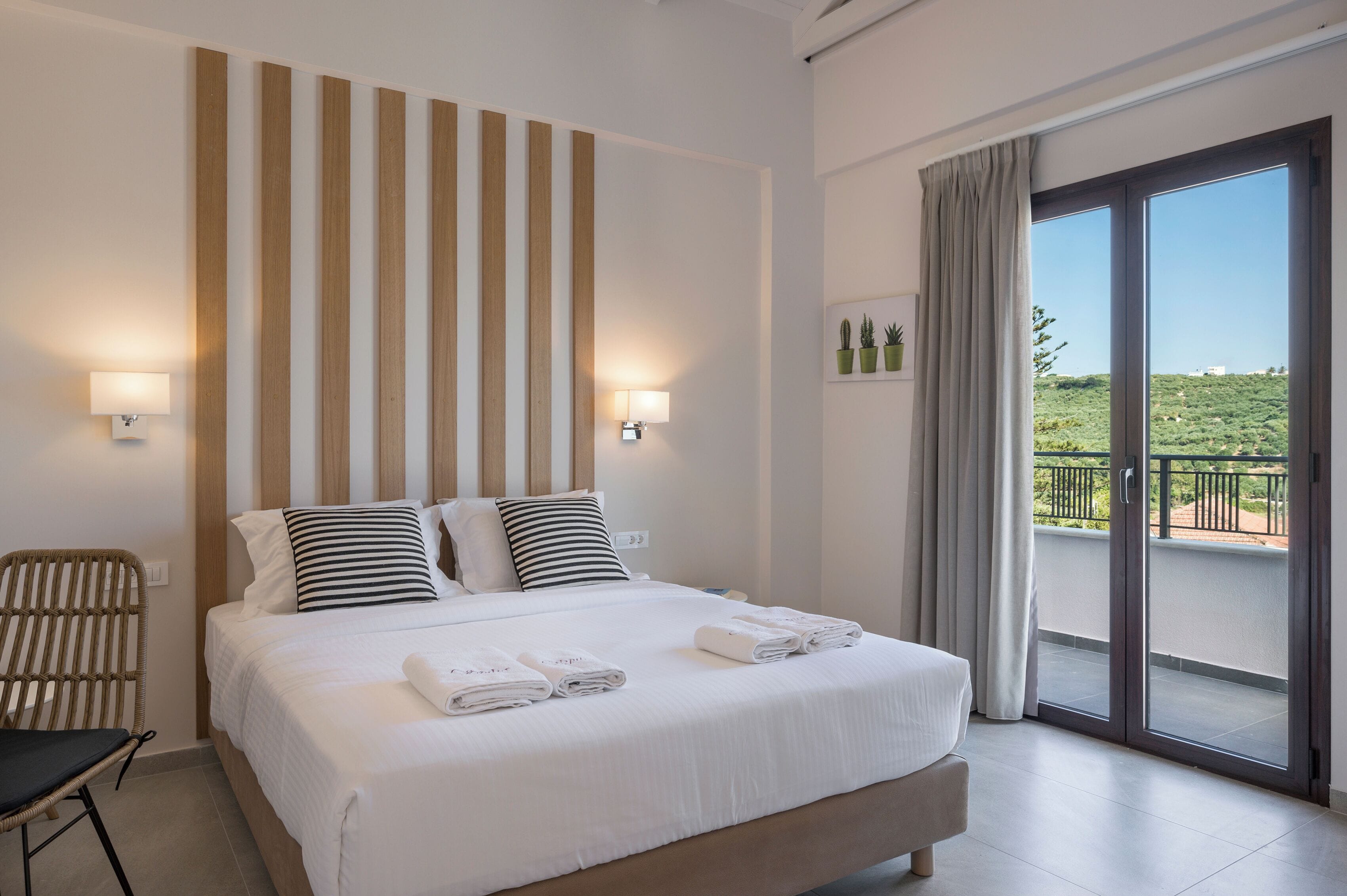 A hotel room with a large bed, two bedside lamps, a view of greenery through a sliding glass door, and a wicker chair.