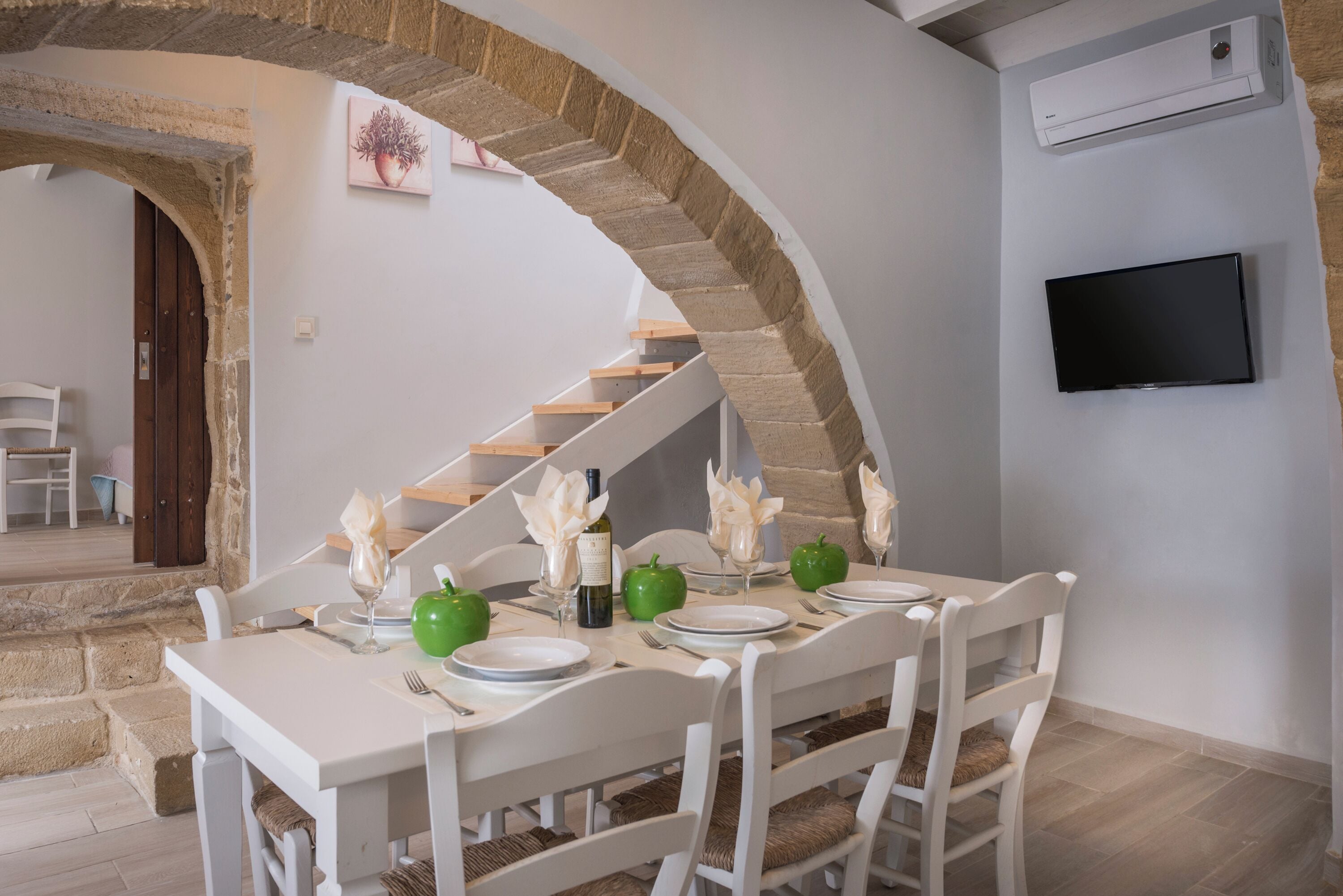 Un comedor con una mesa blanca dispuesta para cuatro personas, una escalera de piedra y un televisor de pantalla plana montado en la pared.