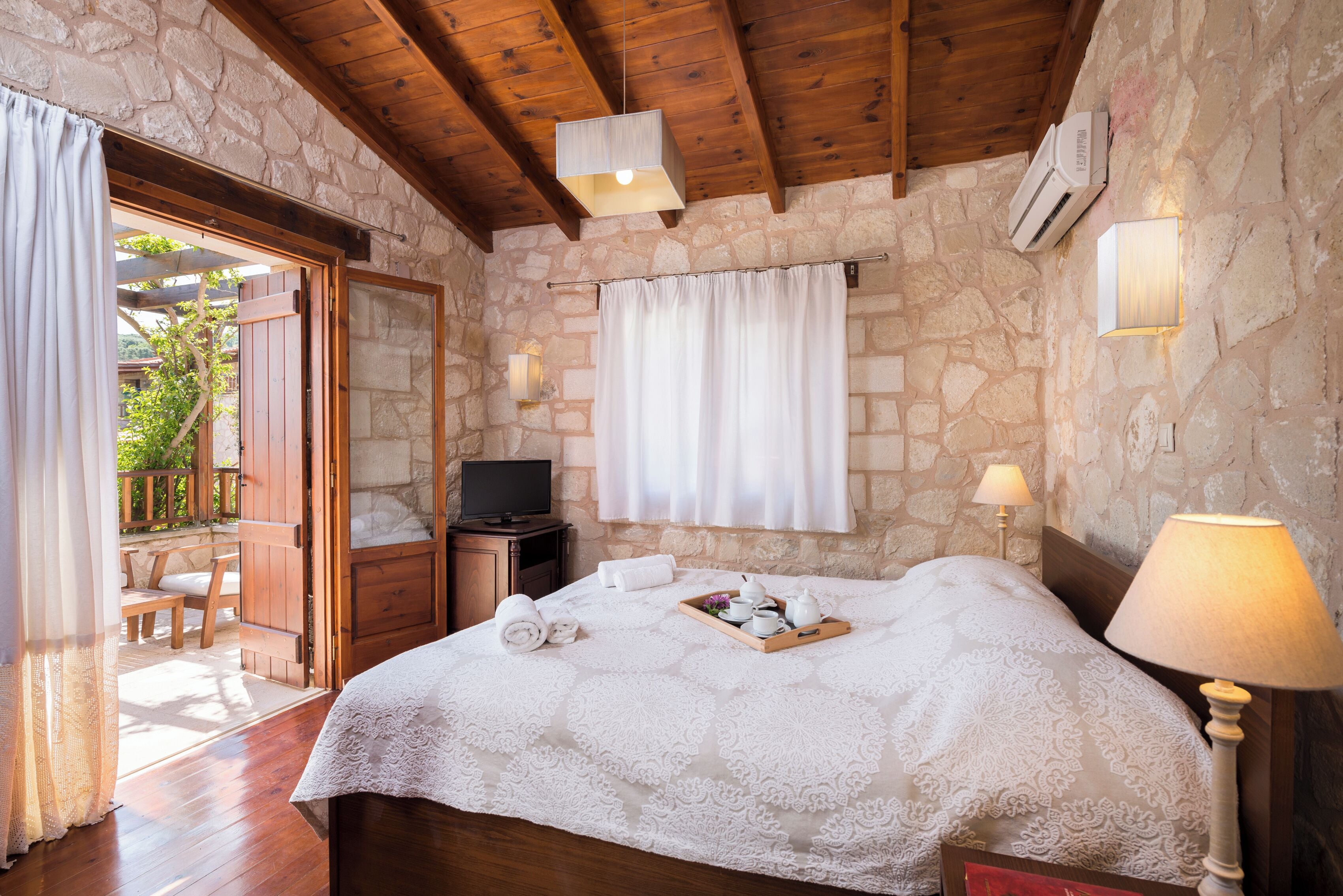 Une chambre avec un mur en pierre, un plafond en bois, un lit recouvert d’une courtepointe blanche, deux lampes de chevet, un téléviseur posé sur un meuble en bois et un plateau contenant des articles de toilette.