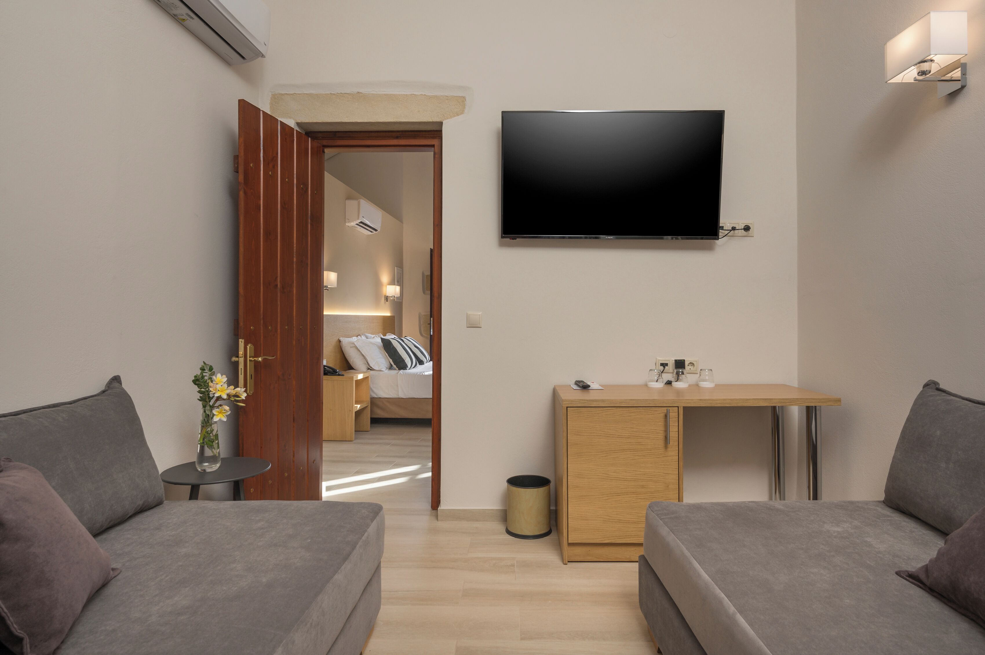 Moderni hotellihuone, jossa on litteänäyttöinen televisio, pieni puinen työpöytä, kaksi harmaata sohvaa ja sängyssä puinen pääty.
